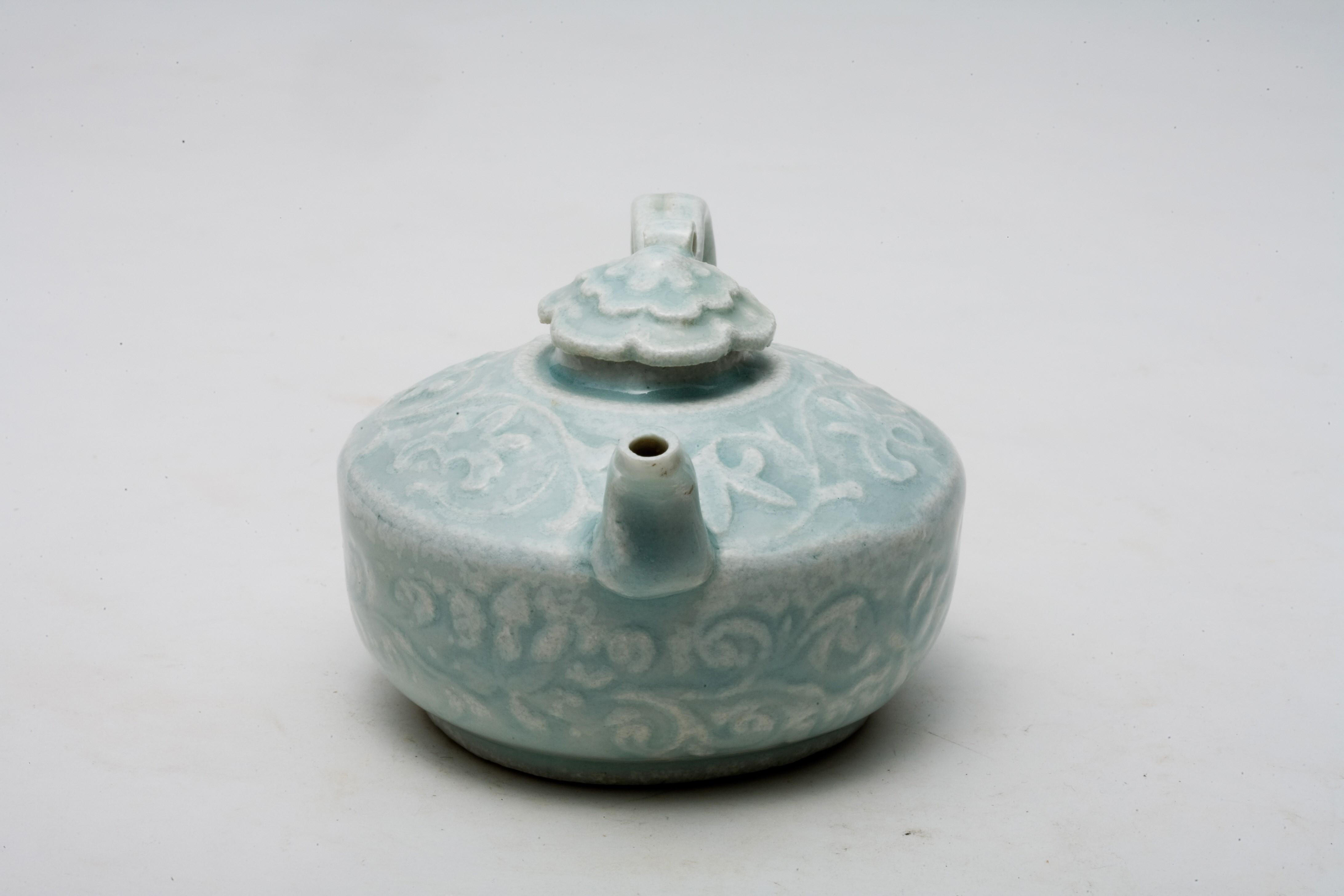 This Qingbai ewer features a rounded body with smooth curves, a short spout, and a looped handle, presenting a harmonious and balanced form. The lid is adorned with a flower-shaped finial, which complements the overall graceful design of the piece.