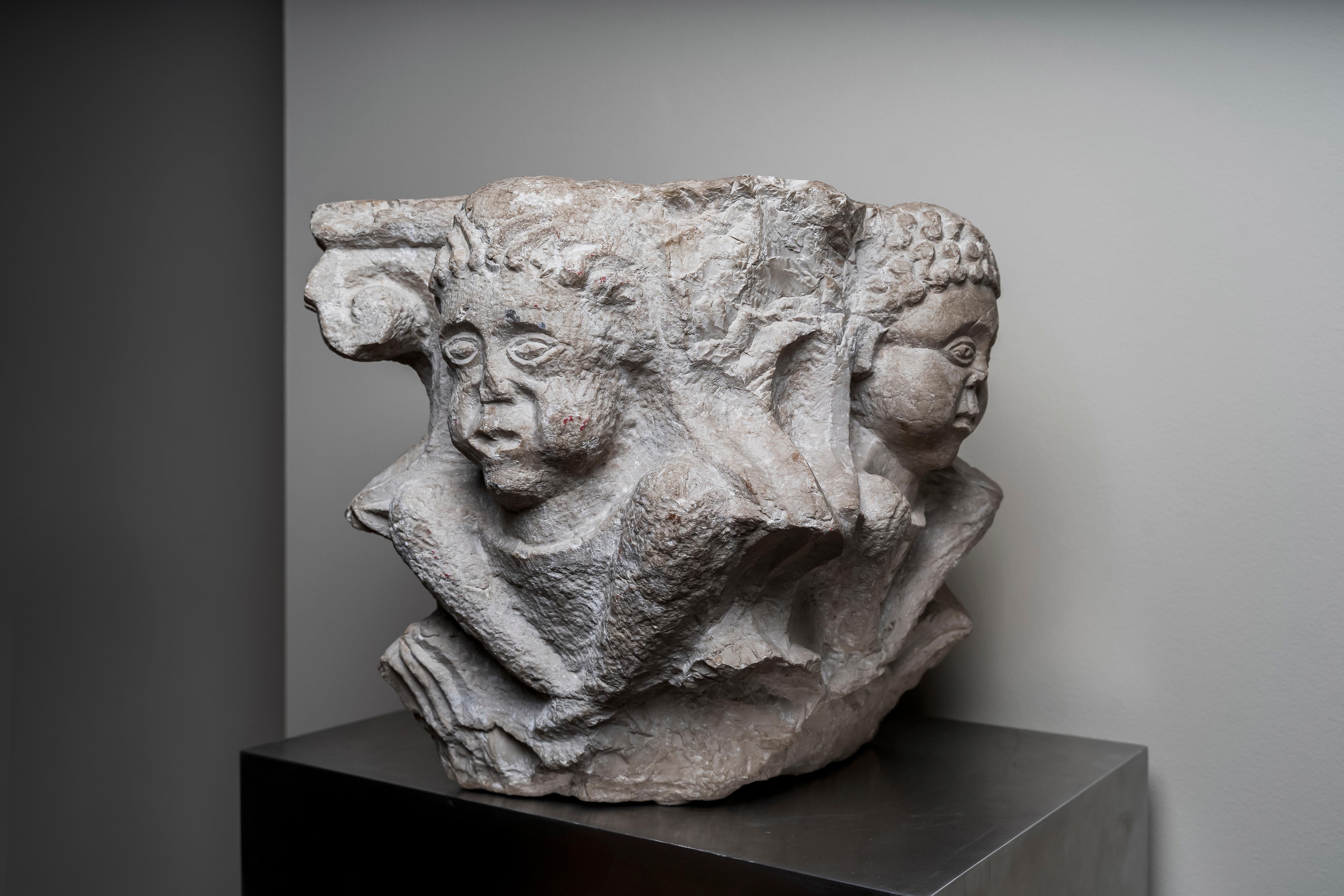 Large stone capital carved on each side in strong relief. The basket is covered with two crowns of vertical acanthus leaves which sprout from the astragal and fill the space between the human heads which decorates each side; each angle is underlined