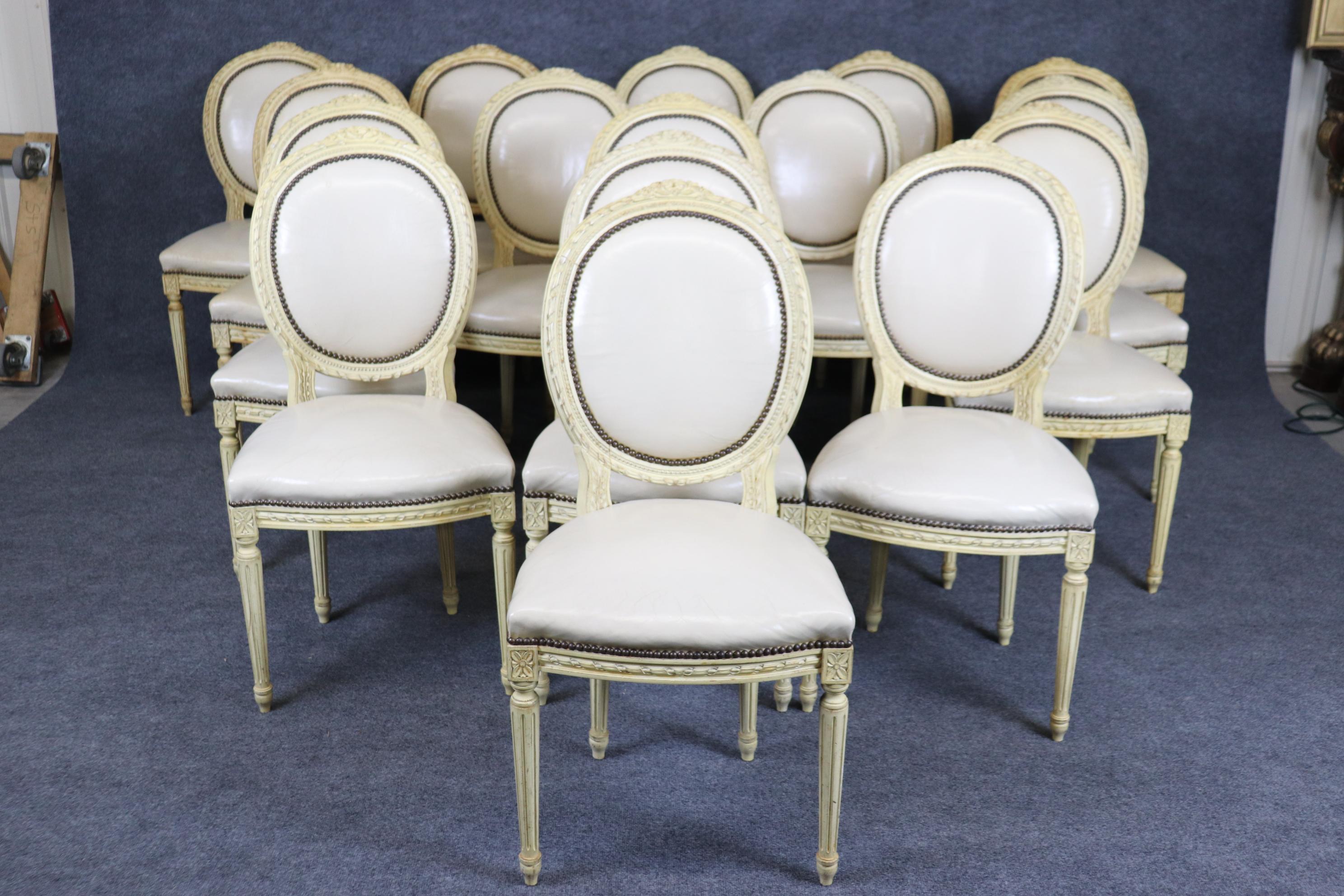 This is a spectacular set of 16 French carved creme painted dining chairs. The chairs in an antique white or creme finish and has a leather or perhaps faux leather upholstery. The upholstery as the chair frames will show signs of use and wear but is