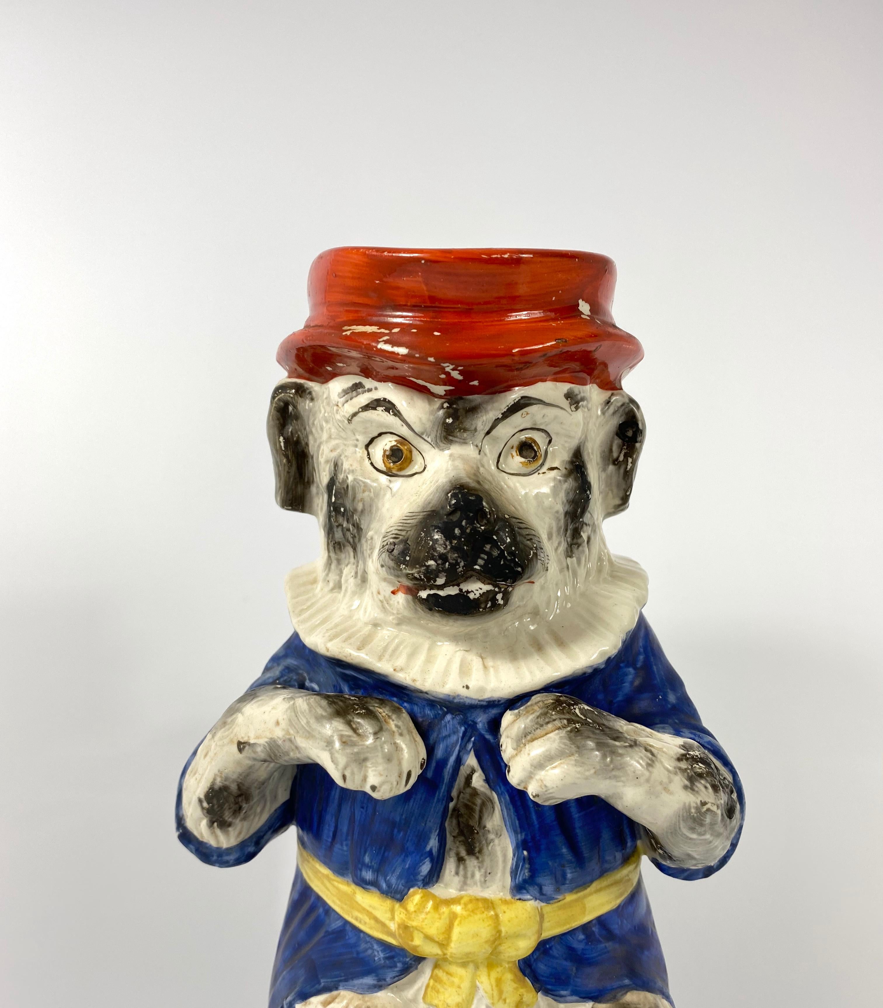 A rare Staffordshire pottery jug, circa 1860. Unusually and amusingly modelled as Mr. Punch’s dog, Toby. The dog stood on its hind legs, wearing a blue coat, a ruff, and his hat forming the spout. Having a simulated rope twist handle, and set upon a