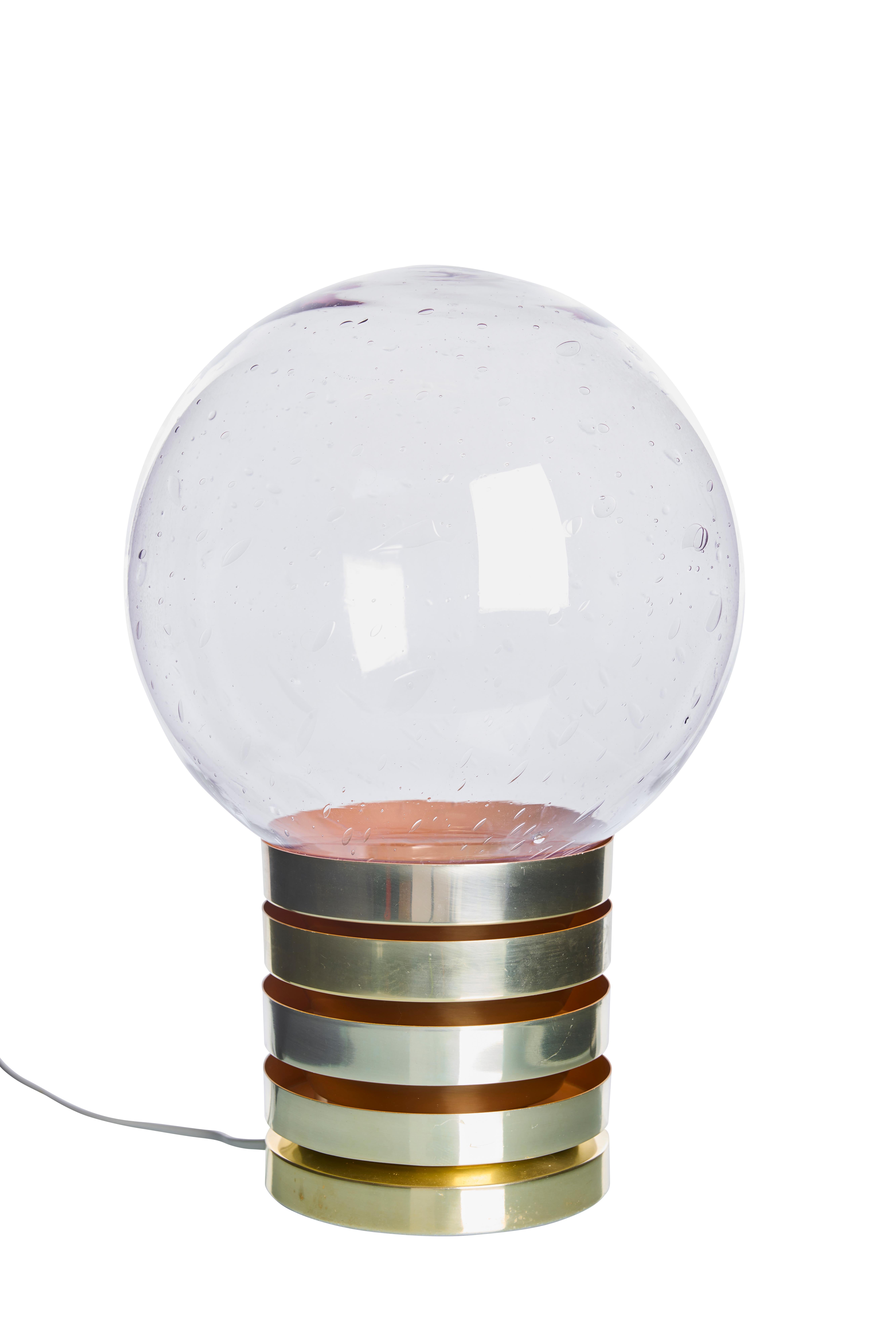 La lampe présente un design distinctif avec une série d'anneaux en laiton poli encerclant sa base. Le laiton chaud est magnifiquement complété par un accent orange intérieur qui ajoute une touche d'élégance moderne. L'intérieur orange vibrant ne