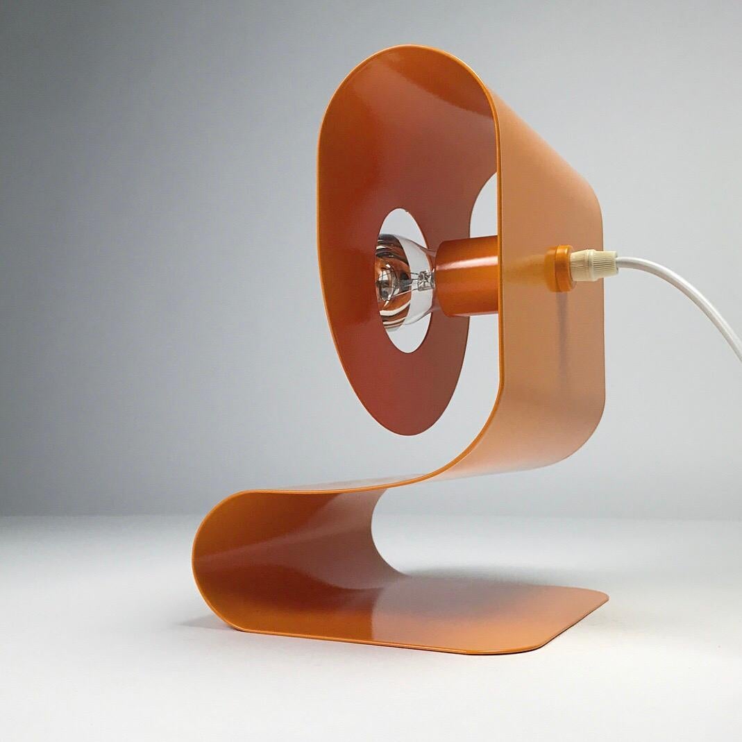 Sometimes a simple idea turns into the ultimate design: An orange lacquered piece of metal bend into a space age shape with a bulb holder and voila! A contemporary table lamp is born. 

This space age table lamp by Sölken was designed in Germany