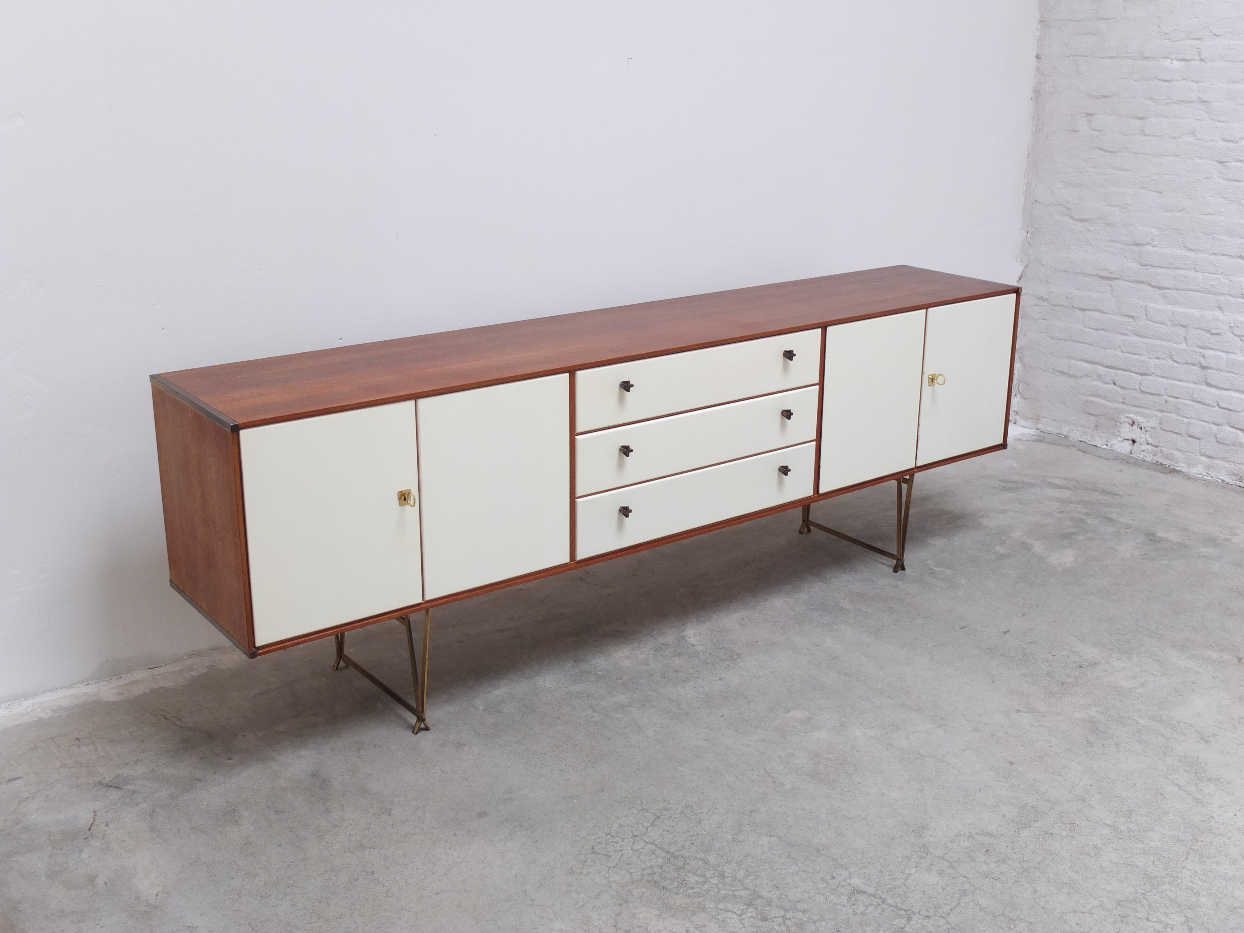 Super refined and elegant sideboard designed by William Watting for Fristho in the 1950s. The combination of teak with brass details all over and the cream colored doors make this a very unique and decorative piece. The slim tapered brass legs that