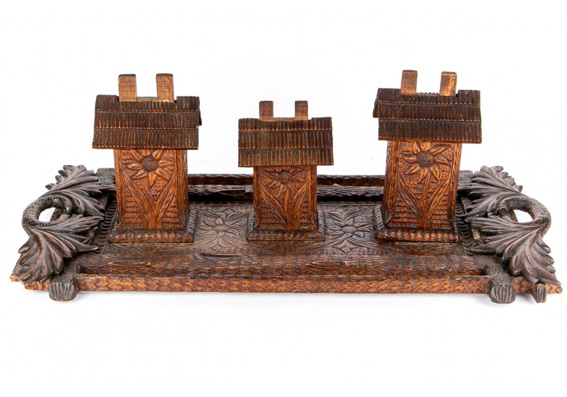 An elaborately carved and stained wood tray densely textured and decorated with sun flowers. Large carved leafy handles on the ends. The tray is set with three house form cigarette dispensers with lift off lids to reveal storage for the cigarettes.
