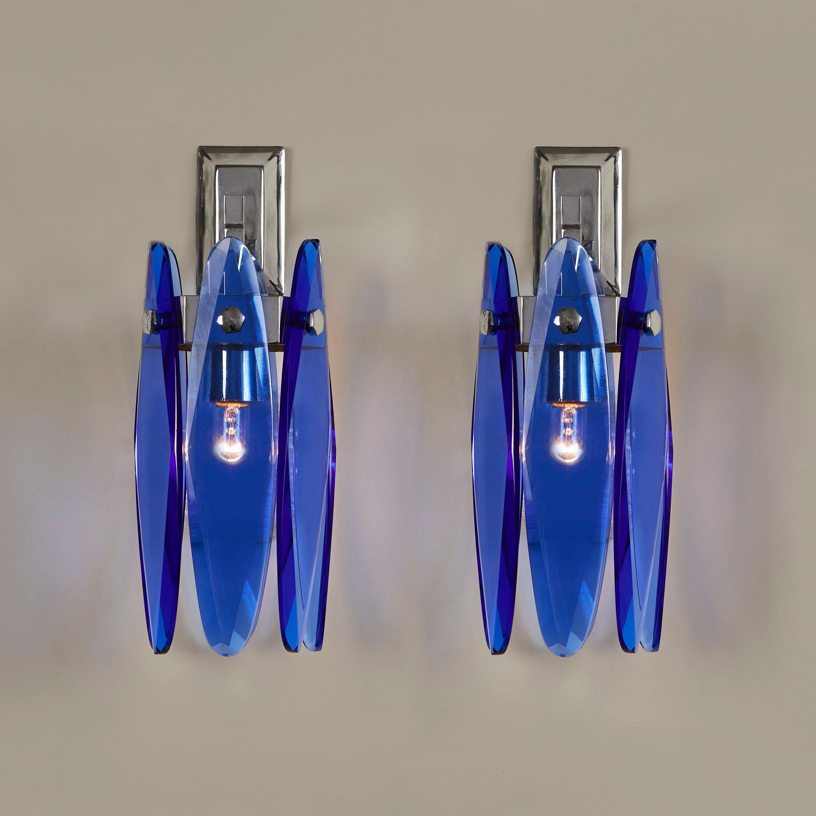 Raros apliques Veca. Cada luz consta de seis gotas de cristal facetado azul cobalto que cuelgan de un marco circular cromado. Se apoya en la pared mediante un brazo cromado unido a una sencilla placa posterior cromada. 