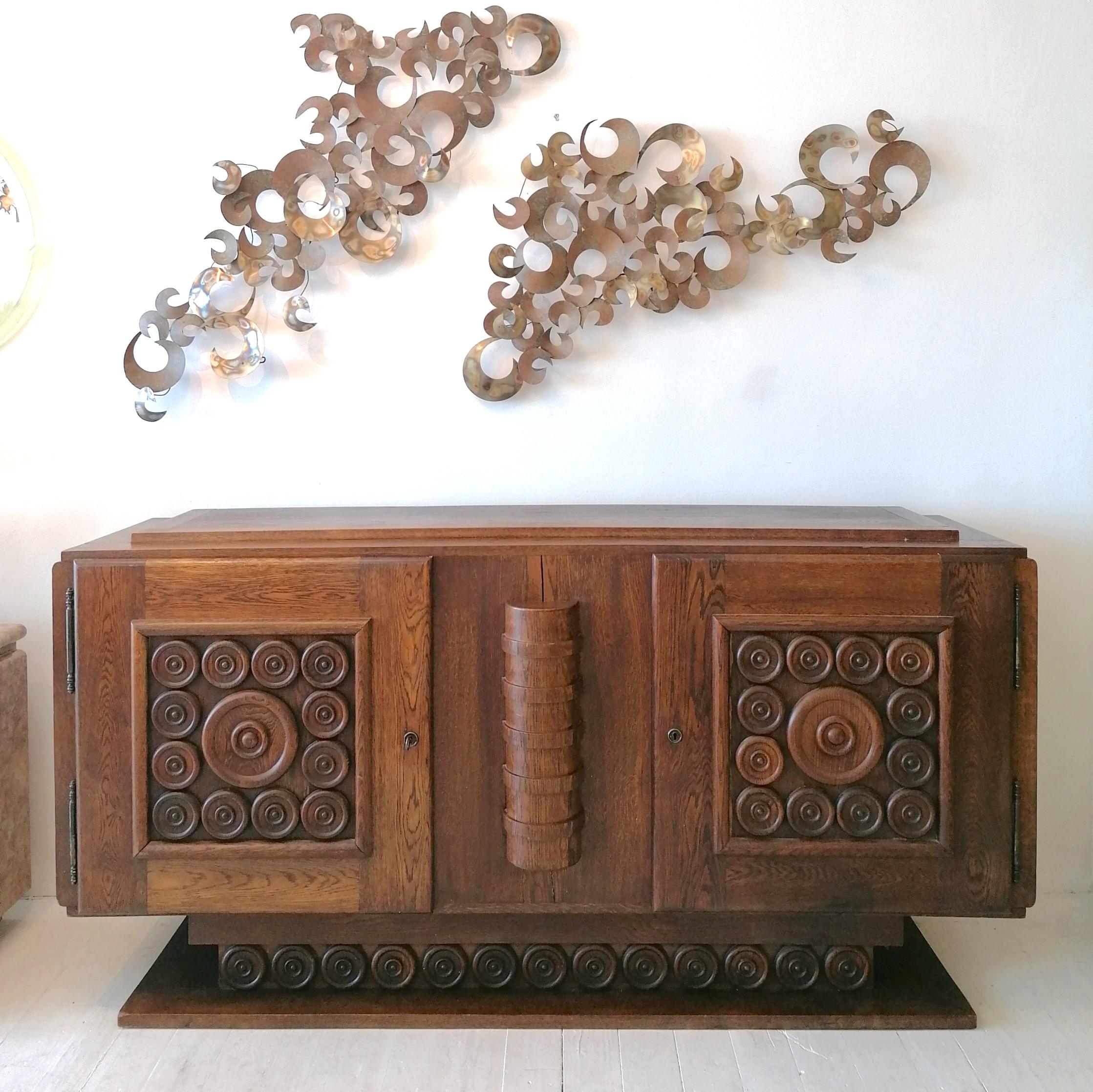 A rare & beautiful Art Deco oak sideboard by Charles Dudouyt, France, circa 1930s, with brutalist styling in the roundel relief design to the front and lower plinth section. Elegant bronzed metal outer hinge details.
It has two locking cabinet