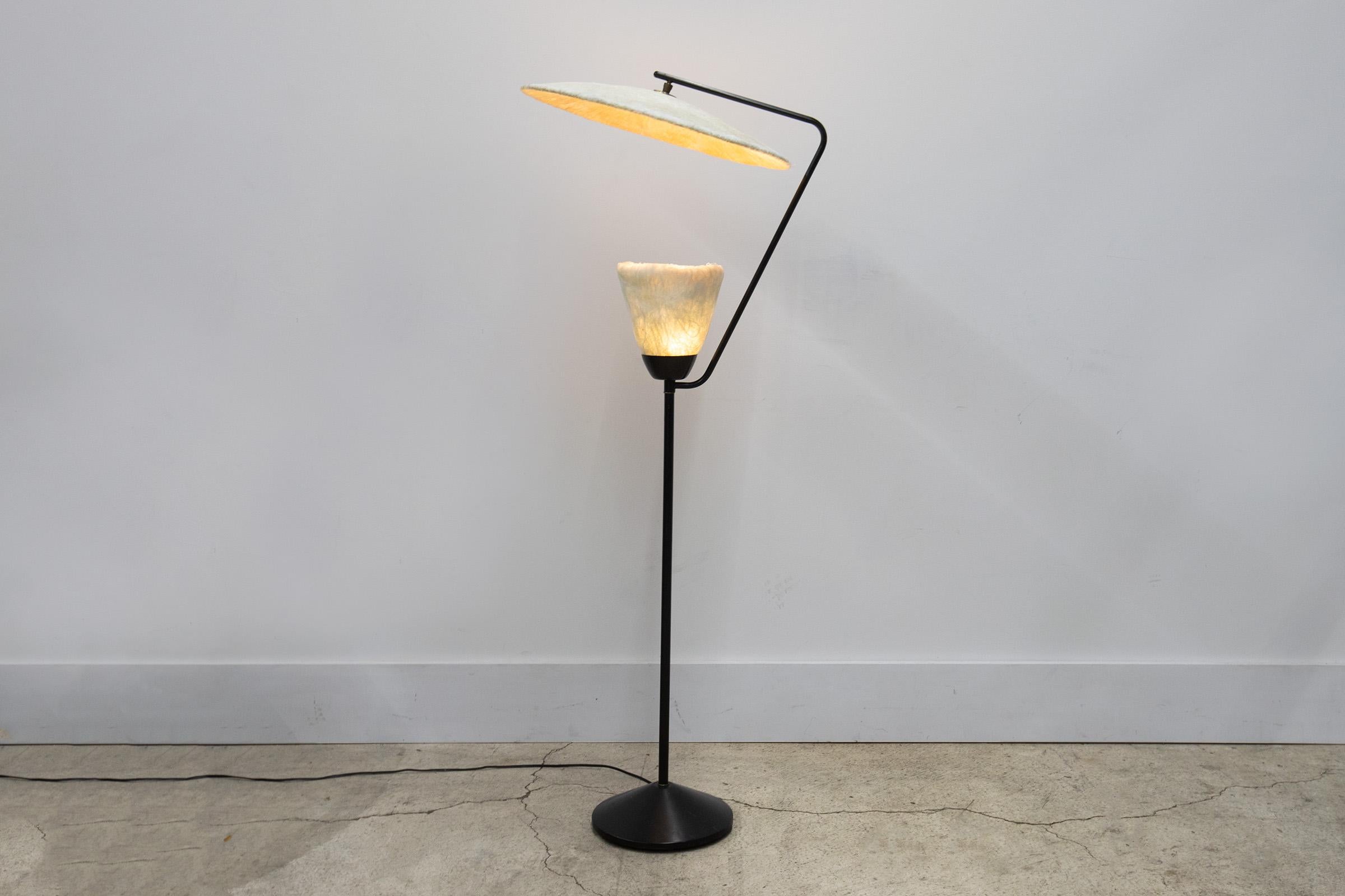 This sophisticated Mid-Century Modern floor lamp was realized in the United States circa 1950. It features a circular button-like base in brass from which two cylindrical black metal supports ascend. One rises from the center straight towards the
