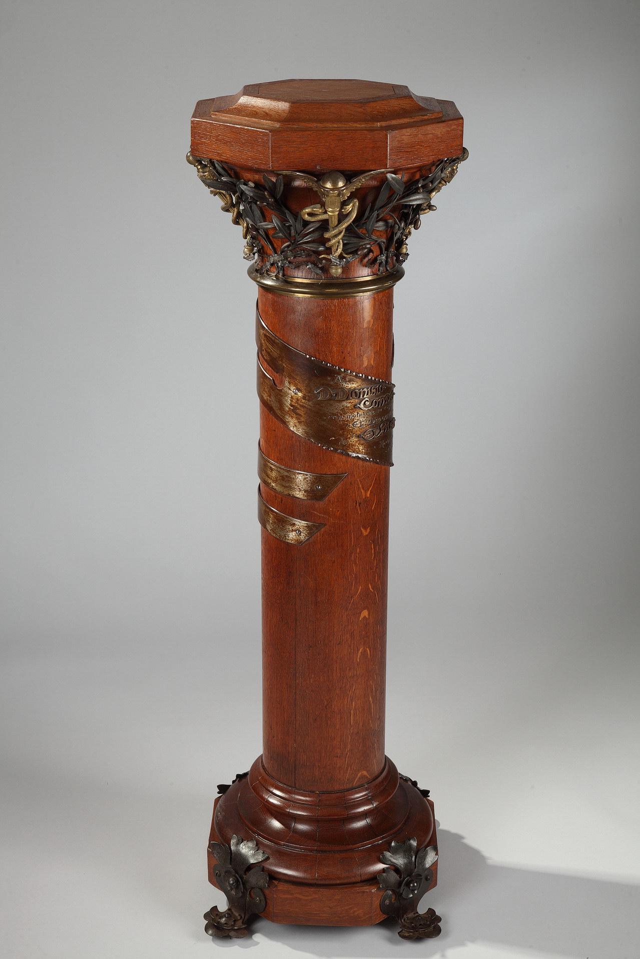 A rare wood column adorned with patinated iron and gilt bronze ornaments. It relies on four bronze feet and is topped with a foliate bronze capital with caduceus. A bronze band is circling the barrel and wear the following dedication, in Spanish: