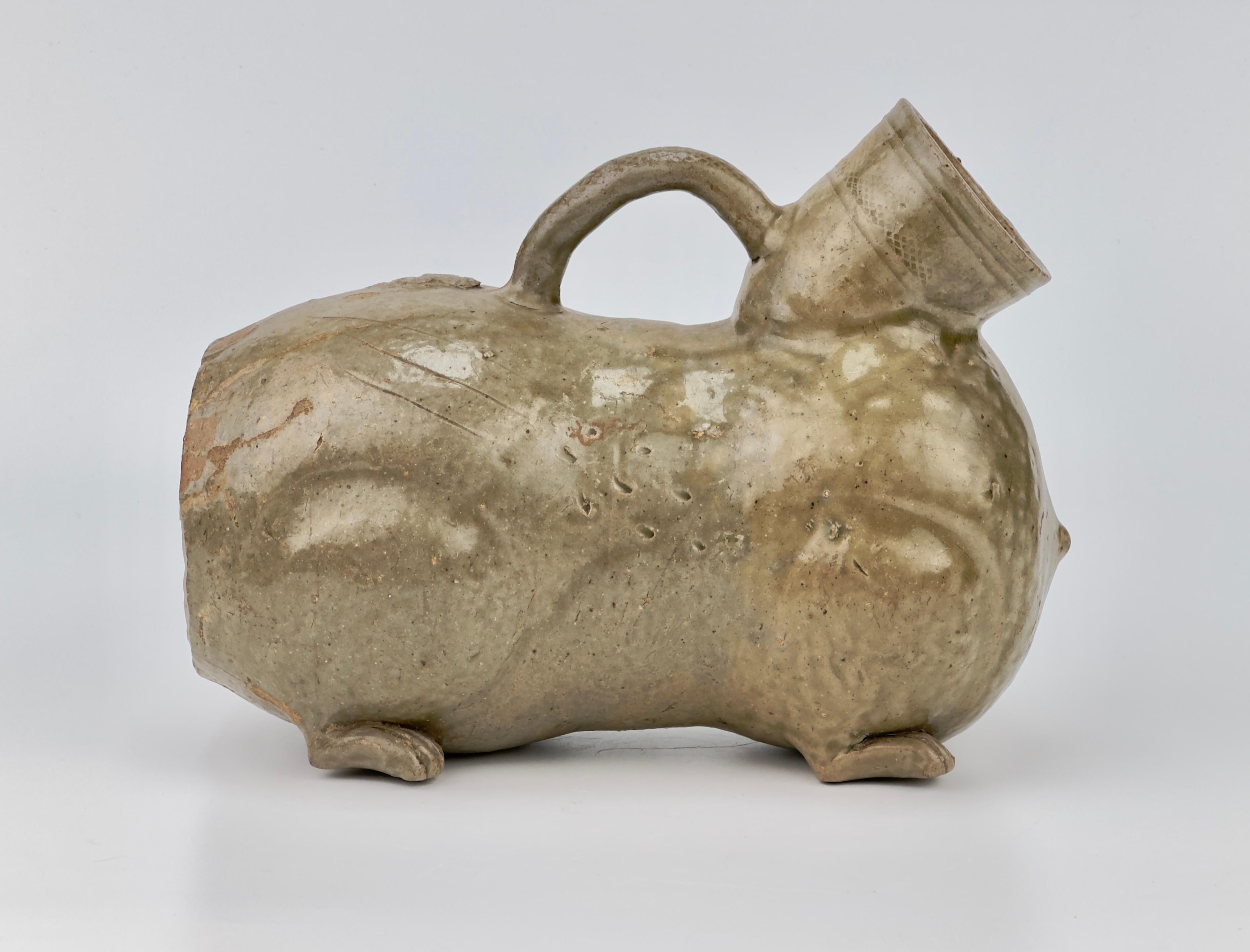 This vessel is well-modeled as a recumbent winged lion with detailed decorative elements. It features a grimacing face with large protruberant eyes under heavy brows and a gaping mouth, which forms a large aperture. The piece has a tail that arches