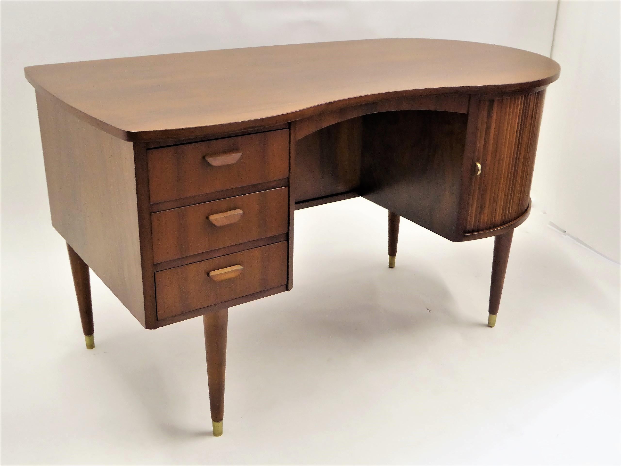 From Denmark, this rarely seen biomorphic double sided teak desk was designed by Gunnar Nielsen Tibergaard. It dates from 1954 and features three drawers on the left with wood pulls. The curved side has two tambour doors that slide and open to