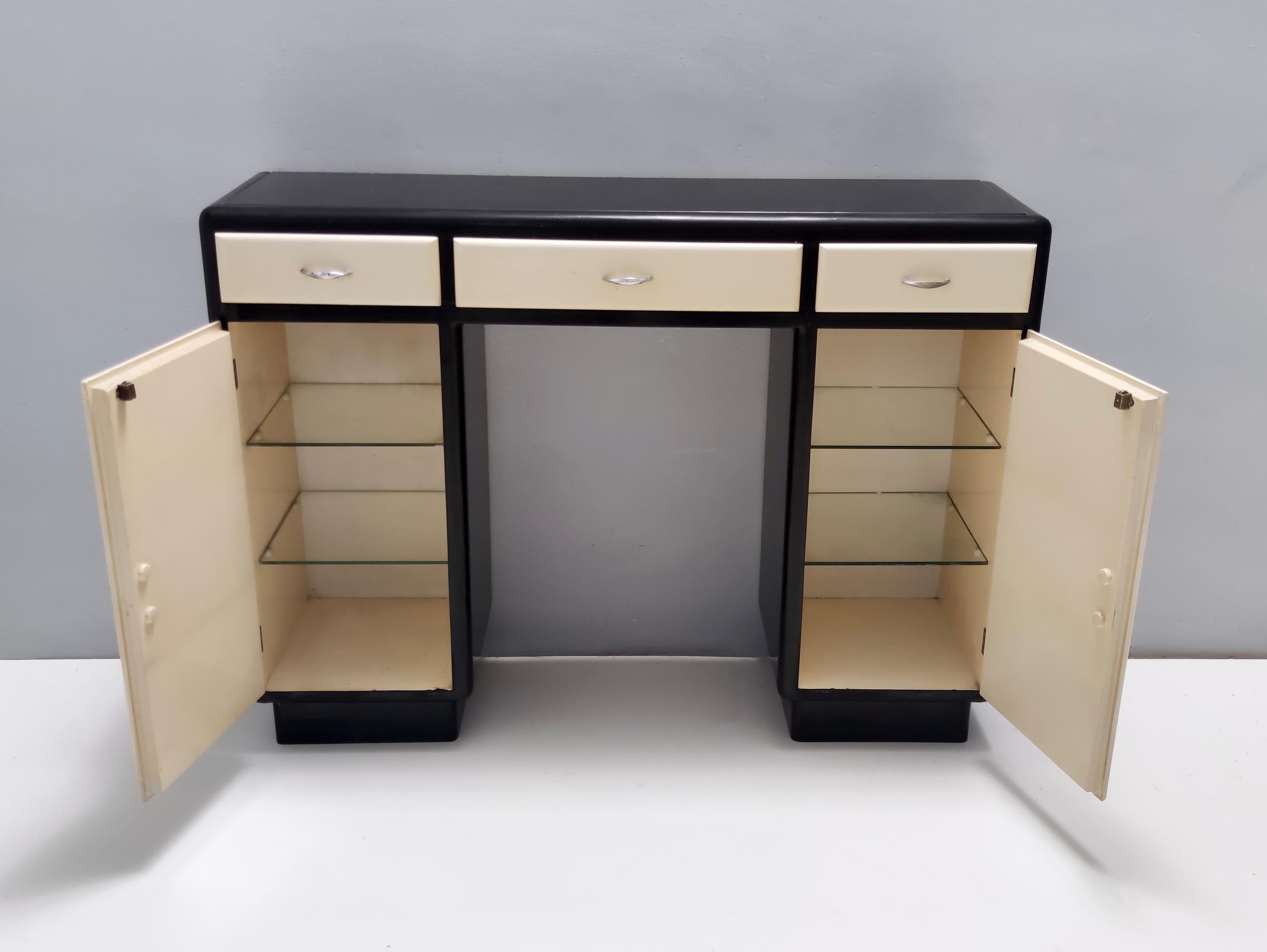 Fabriqué en Italie, années 1940.
Cette console / meuble d'entrée est réalisée en hêtre laqué, avec un plateau en verre opalin noir et présente des poignées en laiton nickelé . 
Il s'agit d'une pièce d'époque, qui peut donc présenter de légères