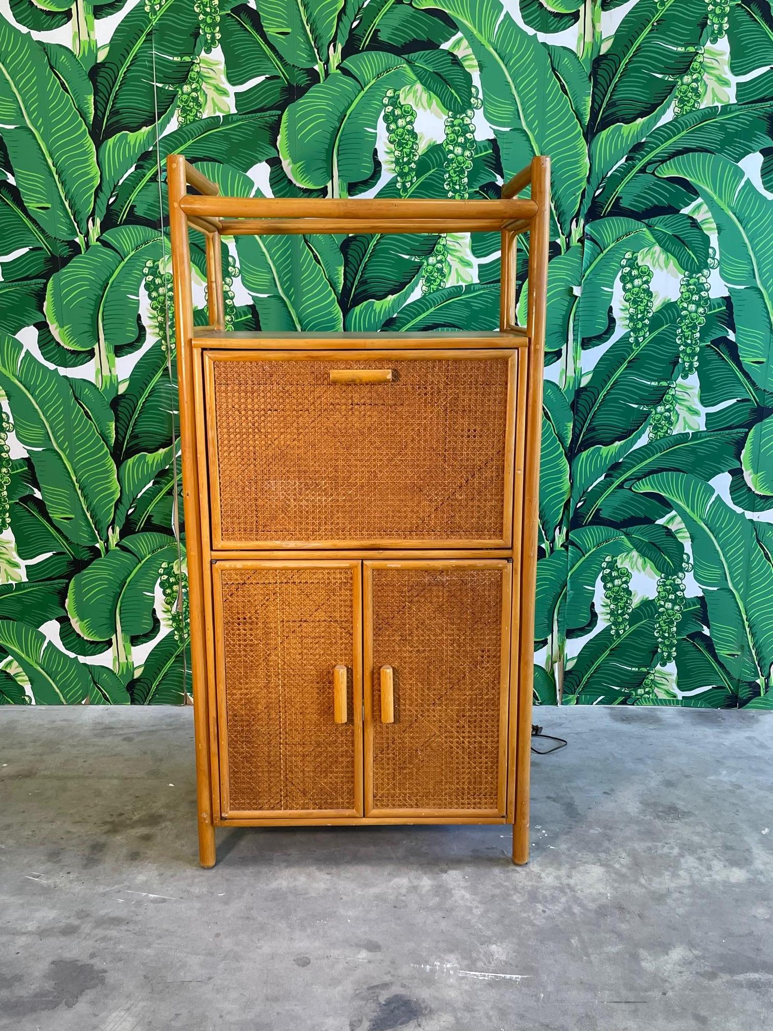 Vintage bar cabinet features wood and rattan construction, drop leaf lighted bar area, and cane veneers. Storage below behind double doors as well as on top shelf. Good vintage condition with imperfections consistent with age. May exhibit scuffs,