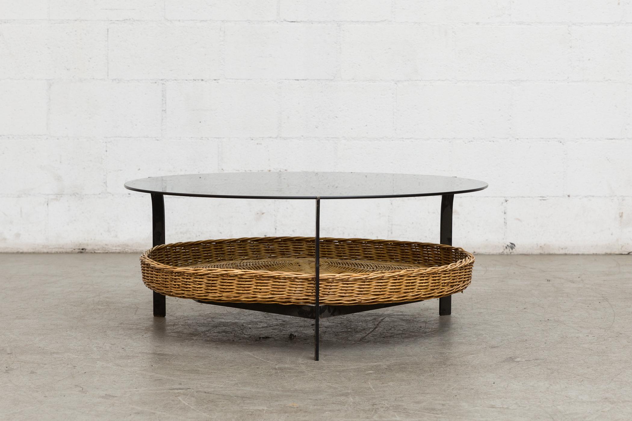 Midcentury round glass coffee table. With woven wicker lower basket black enameled metal frame with slightly turned out detail on metal frame and smoked plate glass. Original condition with visible wear consistent with its age and usage.