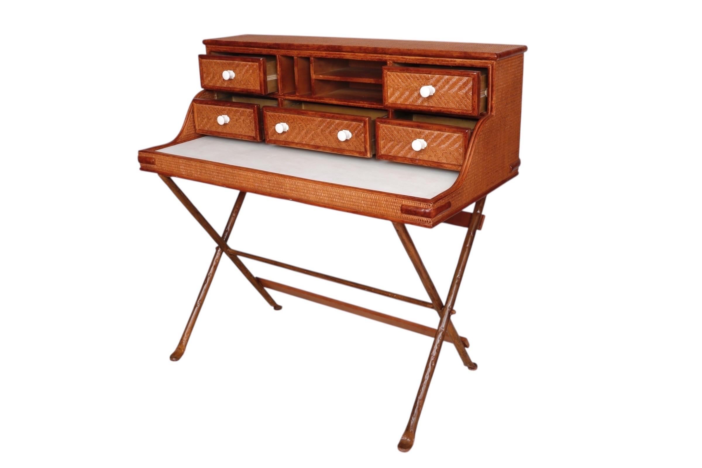 Uno scrittoio in stile Campaigner in rattan. L'area di scrittura è rivestita in ecopelle bianca e il telaio è avvolto in rattan intrecciato. Quattro cassetti nel bureau superiore, che si aprono con maniglie rotonde a pomello bianco, circondano tre