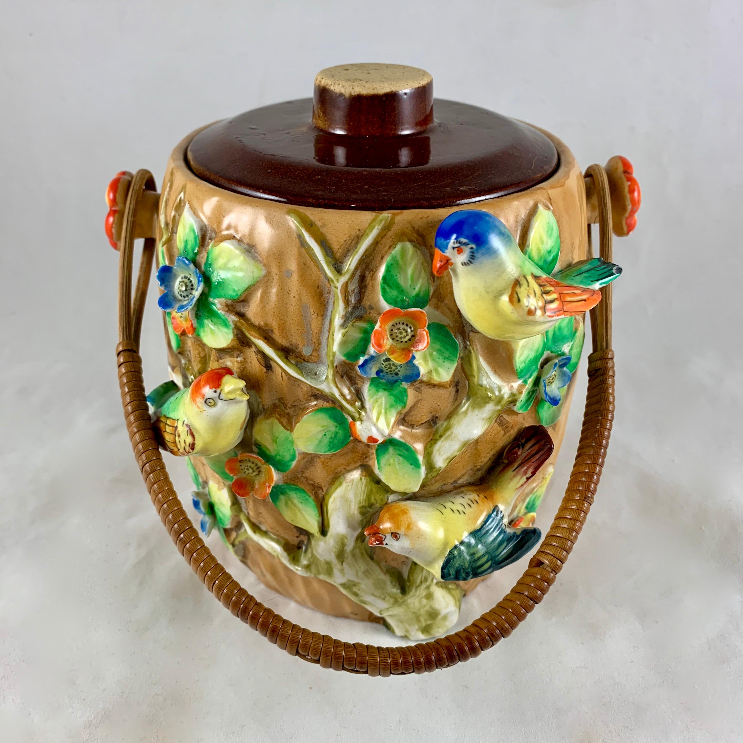 An unusual biscuit barrel marked in red ink – Made in Japan, circa 1920s, Pre WWII.

A highly dimensional barrel showing three birds in high relief perched in a tree surrounded by green leaves and orange and blue blossoms. The birds and flowers