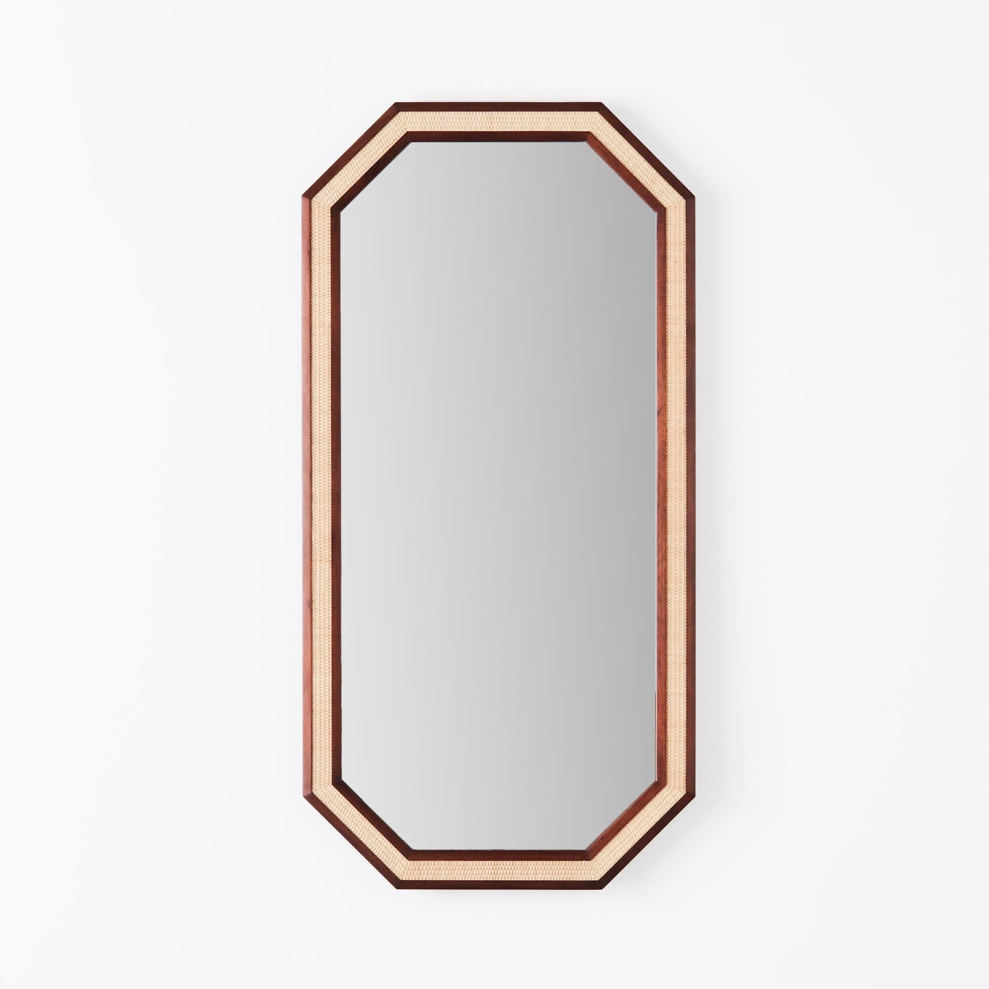 Le Rattan Mirror Large est un miroir de forme octogonale fabriqué en acajou cultivé. Son cadre en bois teinté foncé présente des incrustations de rotin tressé à la main, créant un contraste saisissant et ajoutant de la texture au design. Le bois est