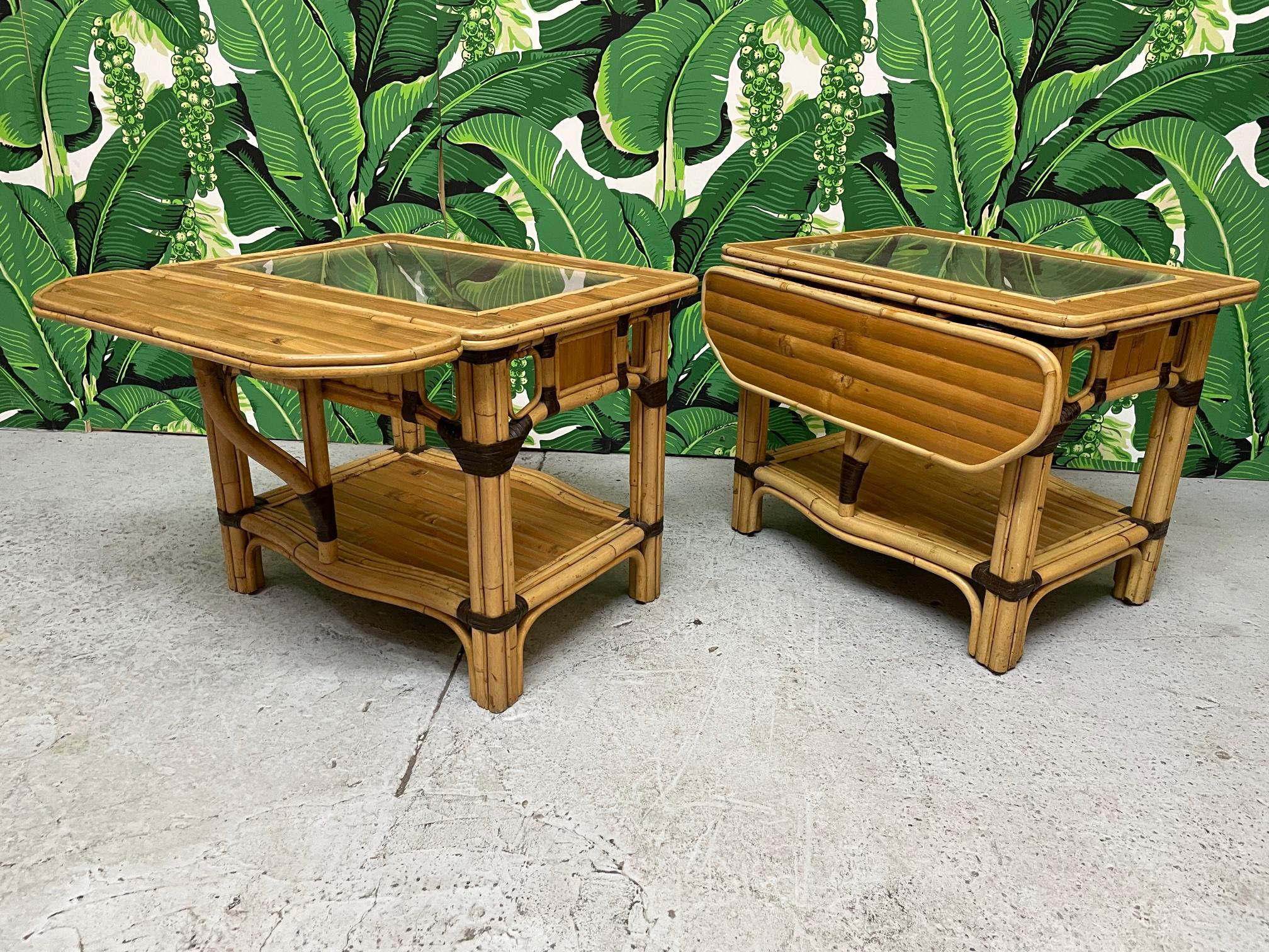 Pair of vintage split reed rattan side tables feature glass tops and unique drop leaf extensions on one side. Good condition with imperfections consistent with age. May exhibit scuffs, marks, or wear, see photos for details. Tops are plexiglass and