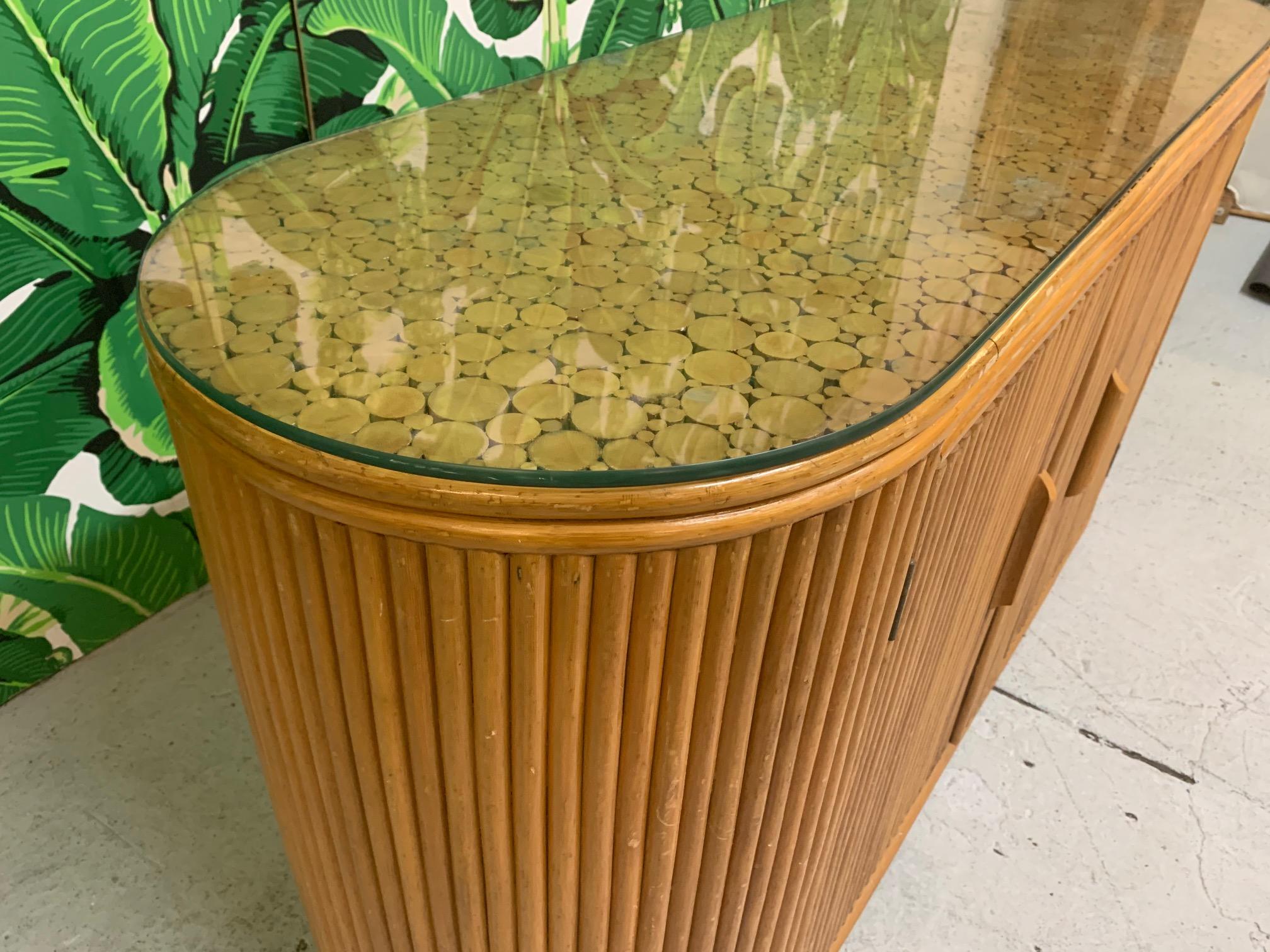 Stunning oval credenza features rattan split reed design with custom cut glass top. Two doors reveal large storage space. Built in the same design aesthetic as the McGuire sheaf of wheat line. Very good condition with only very minor imperfections