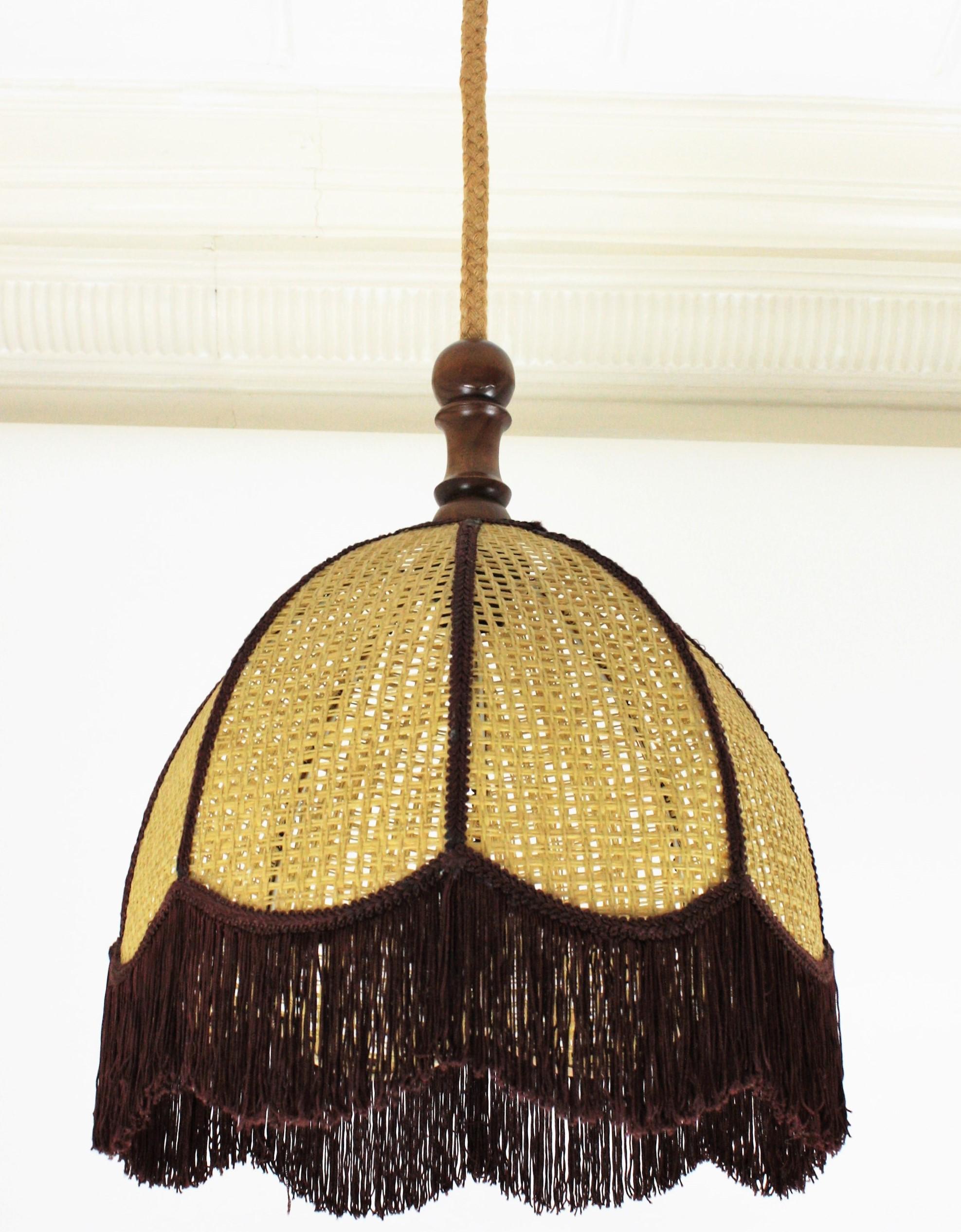 Eye-catching vintage rattan / wicker wire bell shaped lantern or pendant with trims and fringe, Spain, 1960s-1970s.
This midcentury suspension lamp features a wicker wire bell shaped fringe lampshade. It hangs from its original rope and wooden