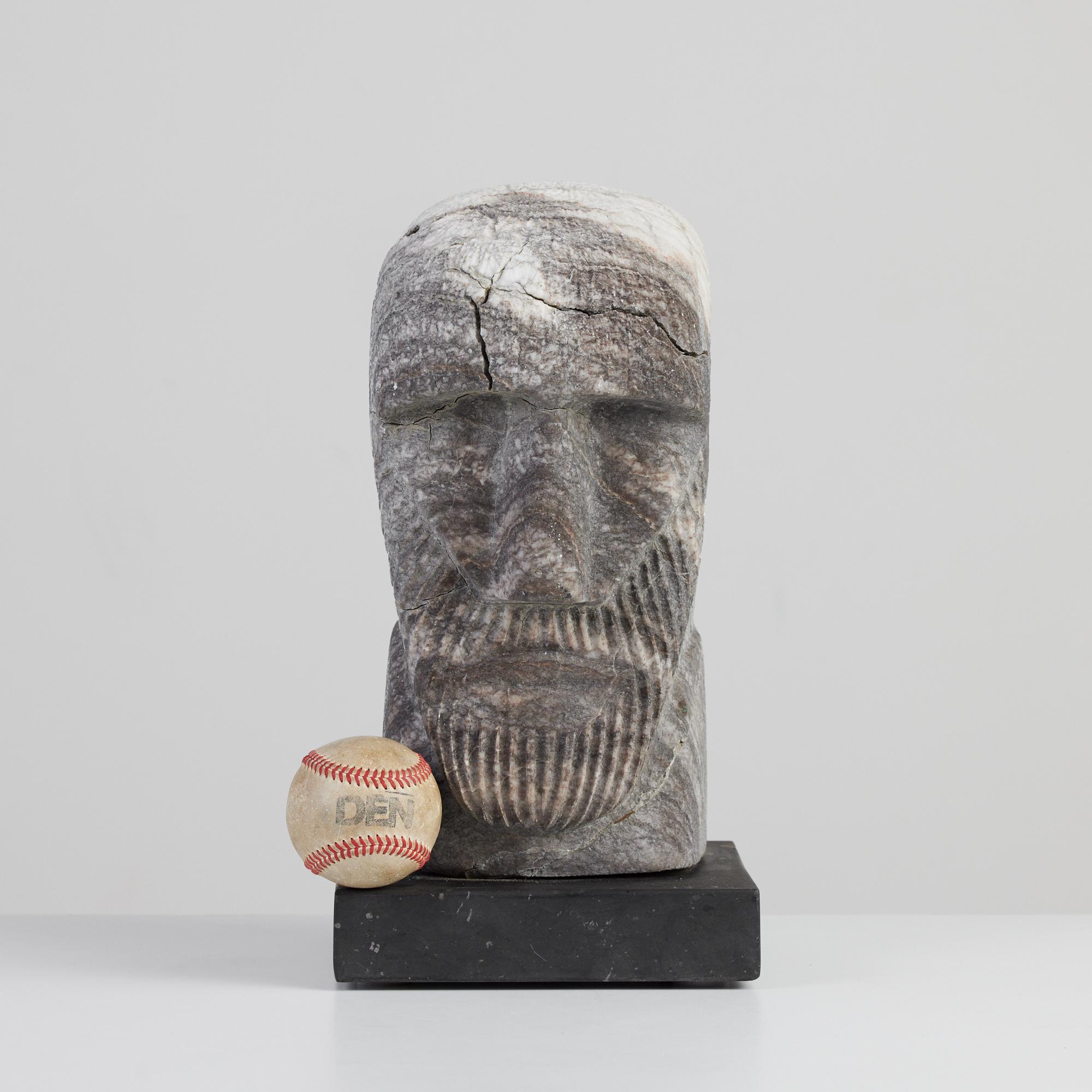 Raw marble figurative carved sculpture with gray and white marbling. This piece appears to be the head of a man with a strong brow, defined nose. and hand carved ribbing around the face to resemble a beard. The sculpture sits atop a polished black