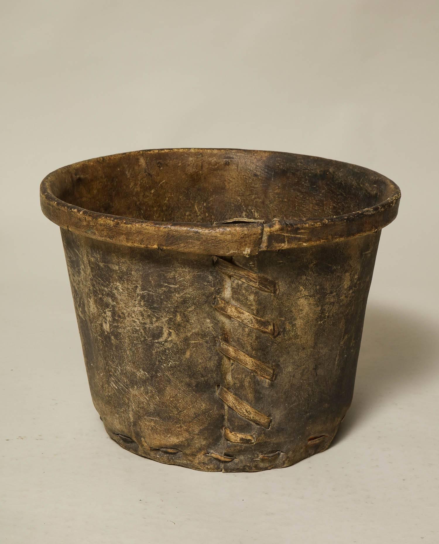 A sturdy and rustic bucket made of rawhide, with rolled rim and stitched seams. Now useful next to a fireplace for logs or as waste paper basket, English, 19th century.