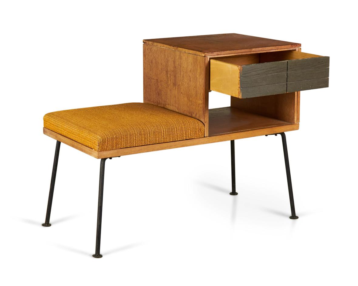 American mid-century walnut telephone bench with a gold textured fabric upholstered seat next to a side table section with a single dark stained, geometrically incised drawer above an open compartment, resting on four angled iron legs with circular