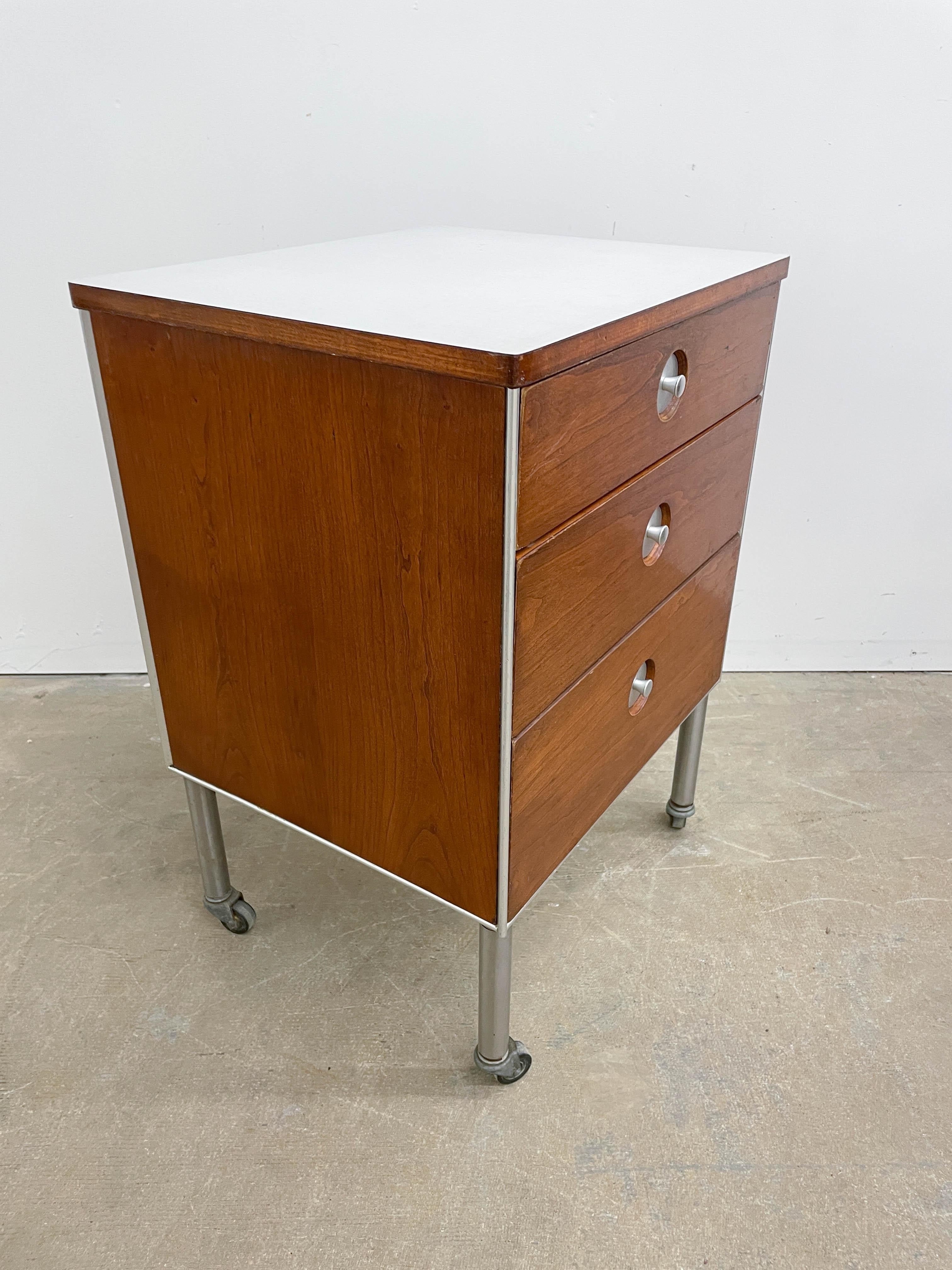 Smartly designed three drawer cabinet bu Raymond Loewy for Hill Rom. Highly functional on swiveling casters, plenty of storage and a high quality formica top. Distinctive formed aluminum pulls, walnut fronted drawers and metal hanging rack on rear.
