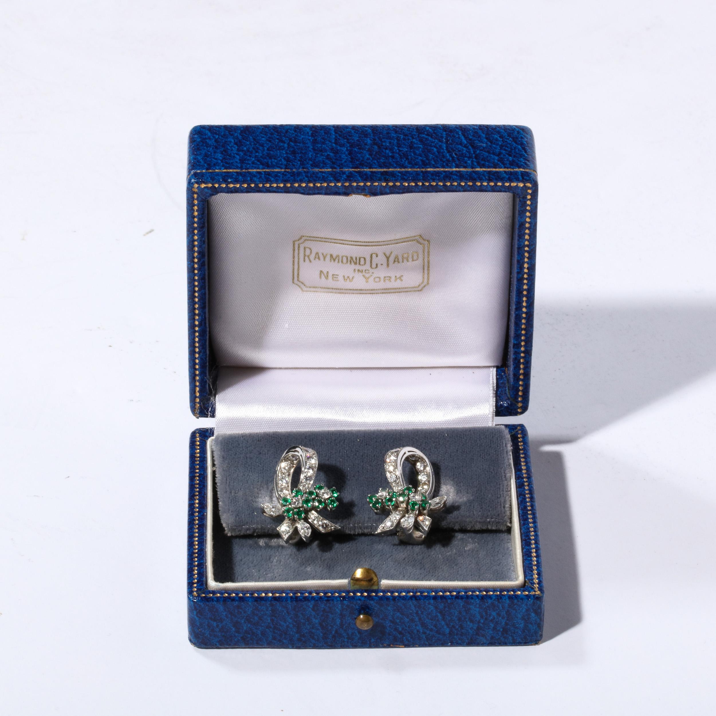 This pair of Raymond Yard platinum and diamond clip on earrings are of bow and flower form with diamond ribbons and emerald flowers.  They have 10 full cut round emeralds and 34 full cut diamonds and 2 marquis cut diamonds . Both marked 
