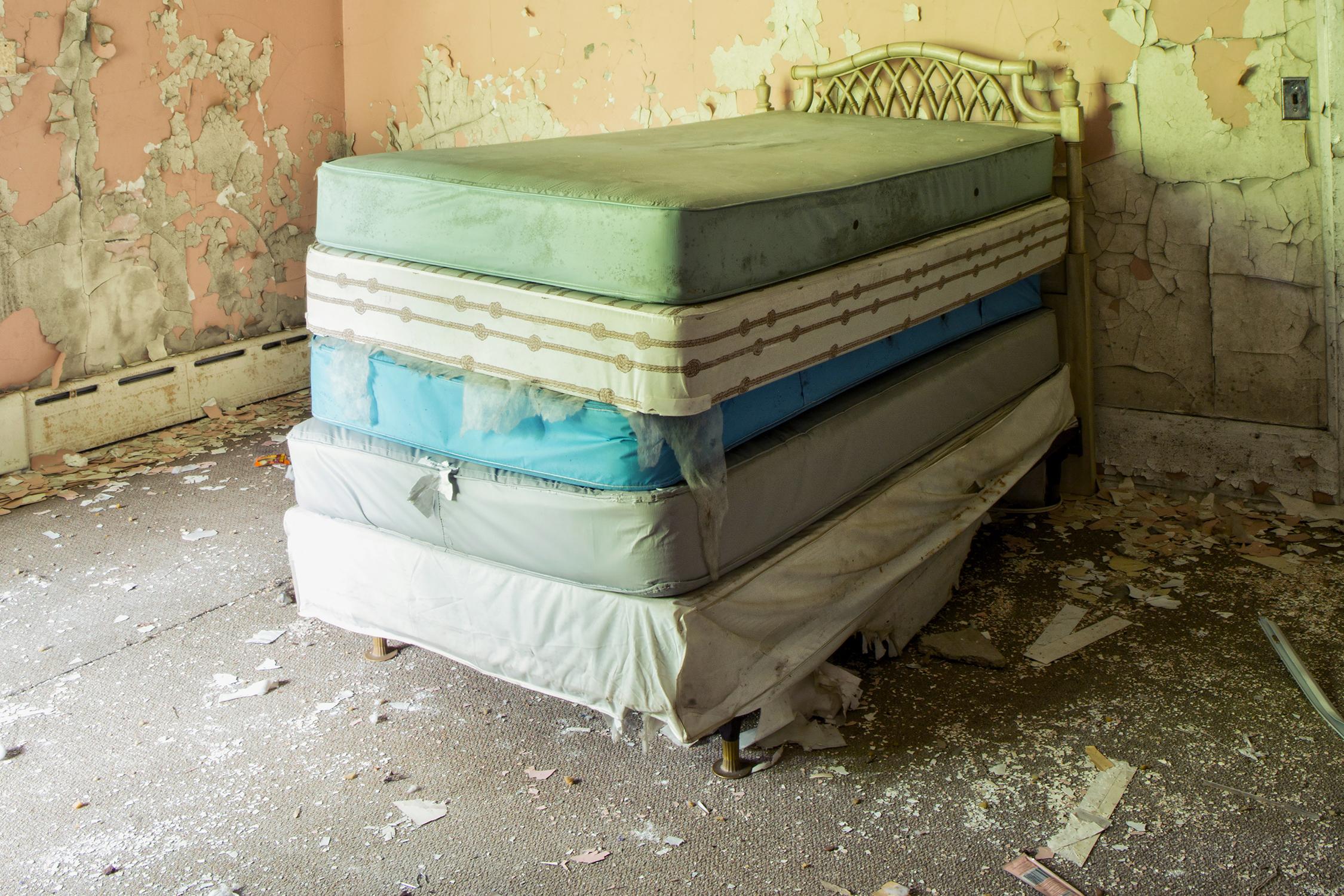 Rebecca Skinner’s “Dream” is a 18 x 12 inch color photograph of a stack of old mattresses stacked on top of each other in the interior of an abandoned building. Peeling paint in peach, yellow and white demonstrate the passing of time. The frameless