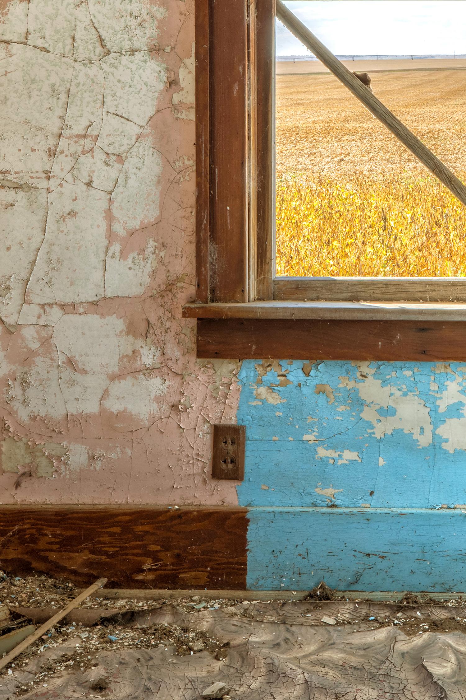 Rebecca Skinner’s “Interior V” is a 18 x 12 inch color photograph taken from inside an abandoned farmhouse in North Dakota. A crooked window in a house in ruin looks out to a field in the beautiful landscape with colors of blue, pink, yellow and