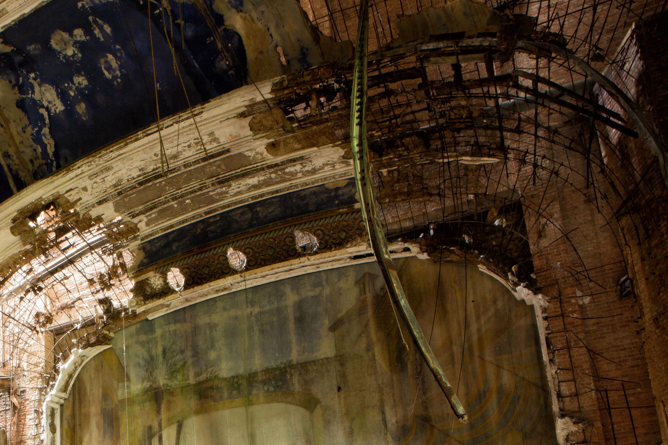 Rebecca Skinner’s “The Palace” is a 16 x 24 inch contemporary color photograph of an abandoned theater in Gary, Indiana. A shredded curtain hangs on the stage in the vast space littered with brick and debris. Light shines through as the ceiling