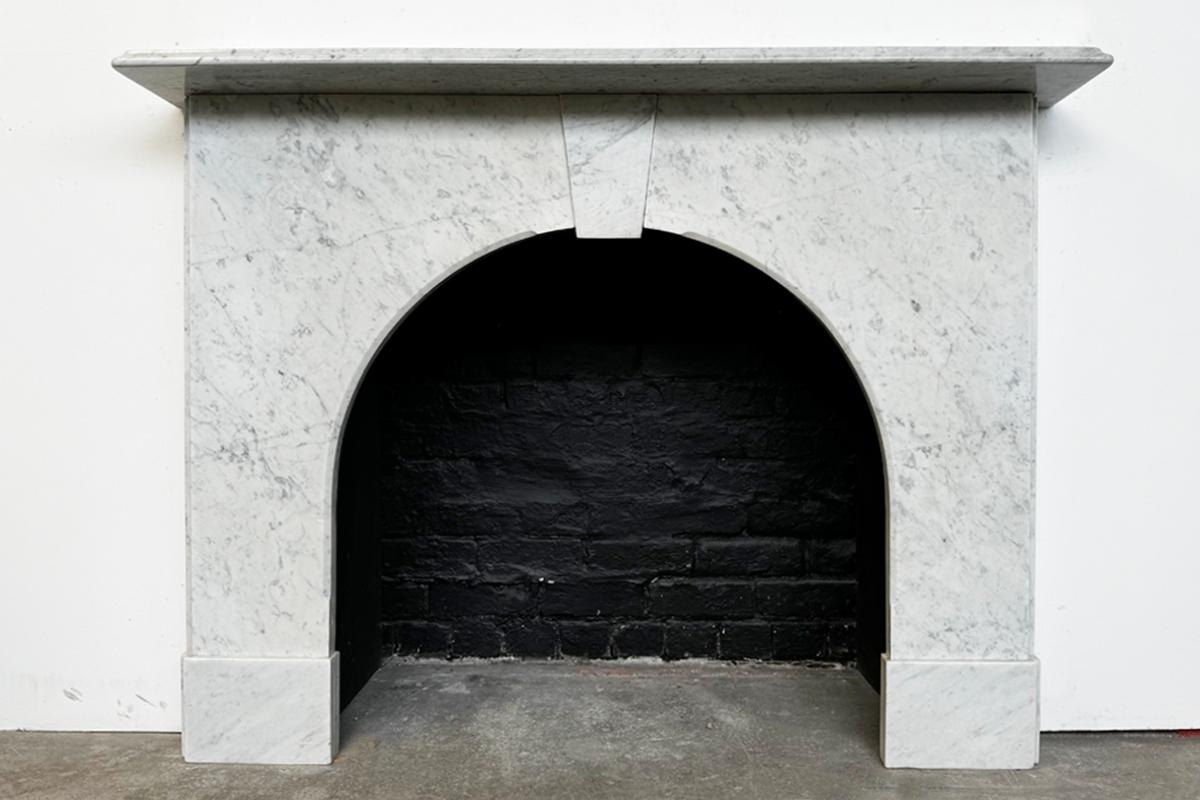 An arched Victorian fireplace surround of good proportions in Carrara marble. The squared edge mantle sits above a plain keystone. The spandrels carved with a four point star within a ring where the Carrara has bush hammered leaving a textured
