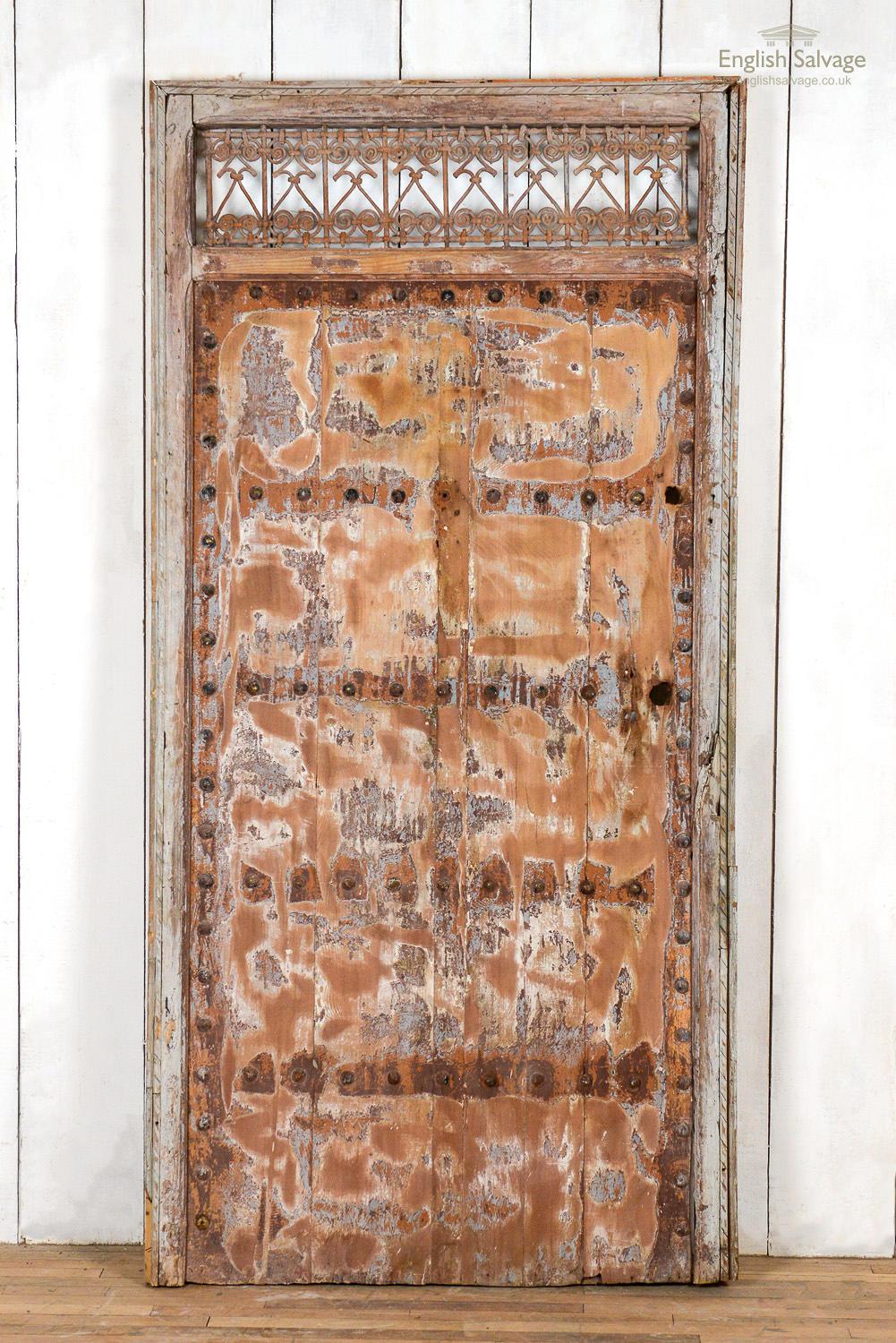 Salvaged sturdy door in frame from Morocco with a weathered patina from paint remnants and age related loss to the wood. The exterior has heavy iron studs/nail heads and the interior has unusual horizontal chamfered panels. Above the door is a