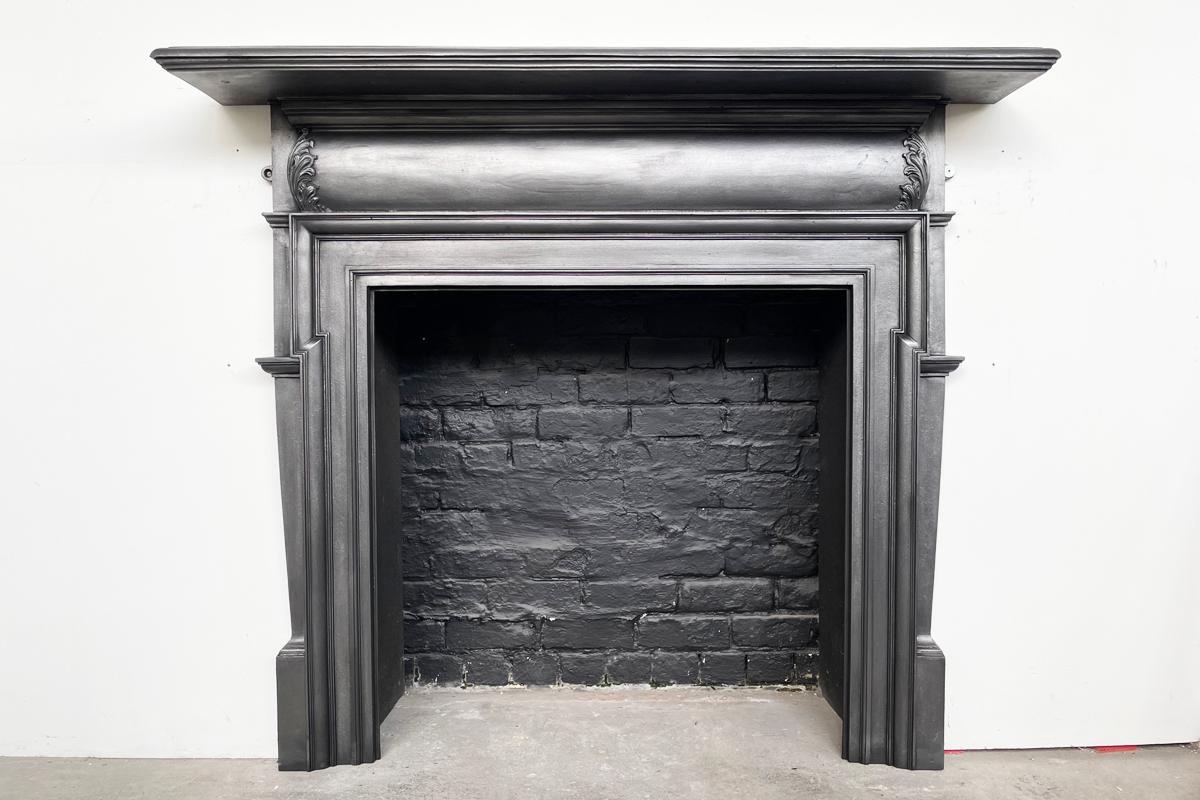 Antique late Victorian cast iron fireplace surround with a cushion frieze terminating in acanthus leaves sits below a generous mantle shelf and above tapering jambs. A dog leg bollection mould frames the aperture. English. Dated 1900.

Fully