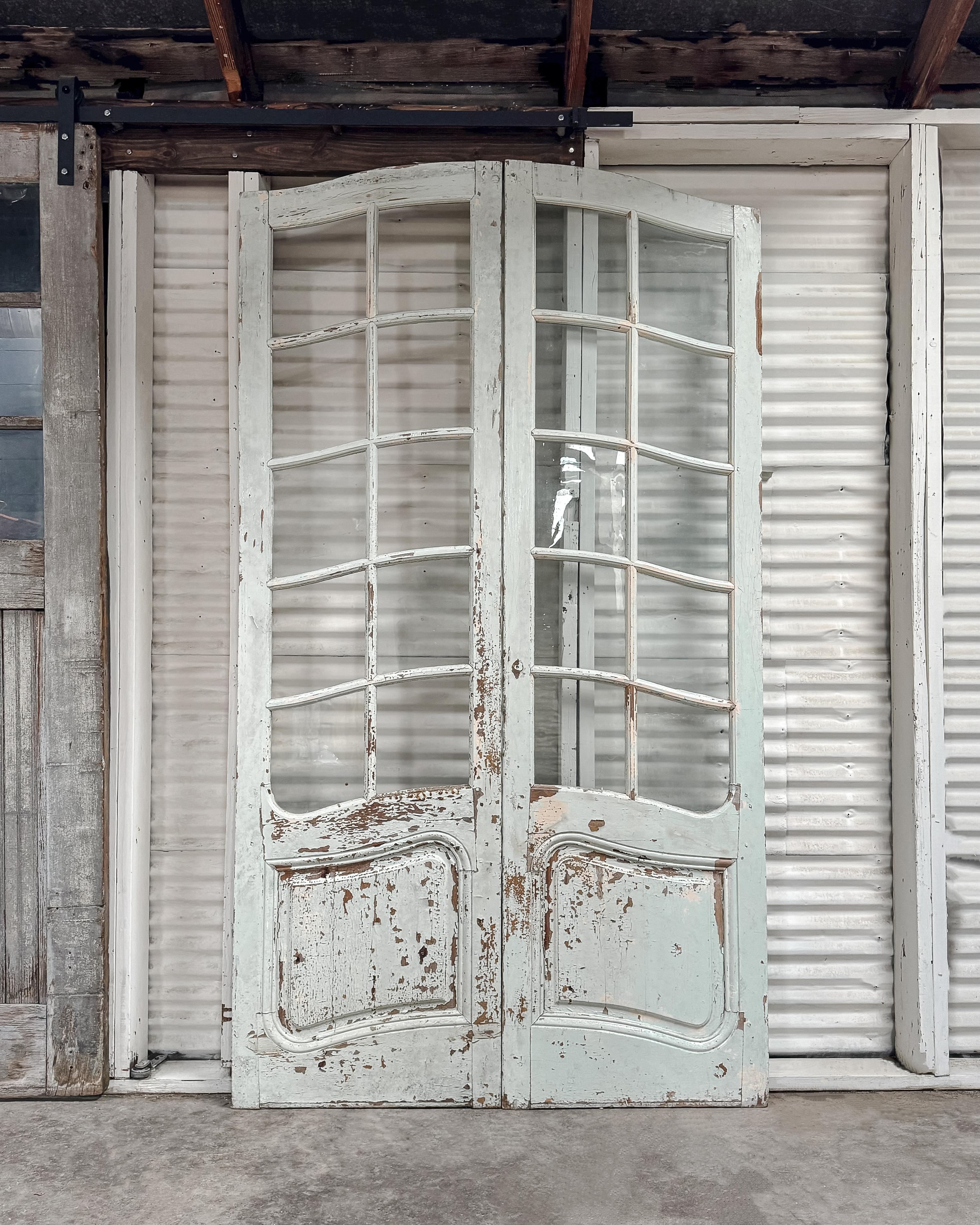 Salvaged provincial-style French exterior doors with a graceful arched top and worn pale seafoam green paint. Each door contains 10 lites that are framed with beveled molding. The lower panels are molded to emulate the door’s curved arch. An iron