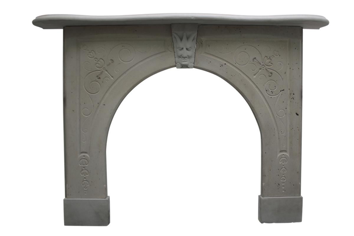 Reclaimed mid-19th century stone fire surround with an arched aperture. Carved detail to the spandrels and a jesters mask to the keystone. Speckled with naturally occurring carbon deposits. Serpentine shelf, circa 1850.

Pictured with an original