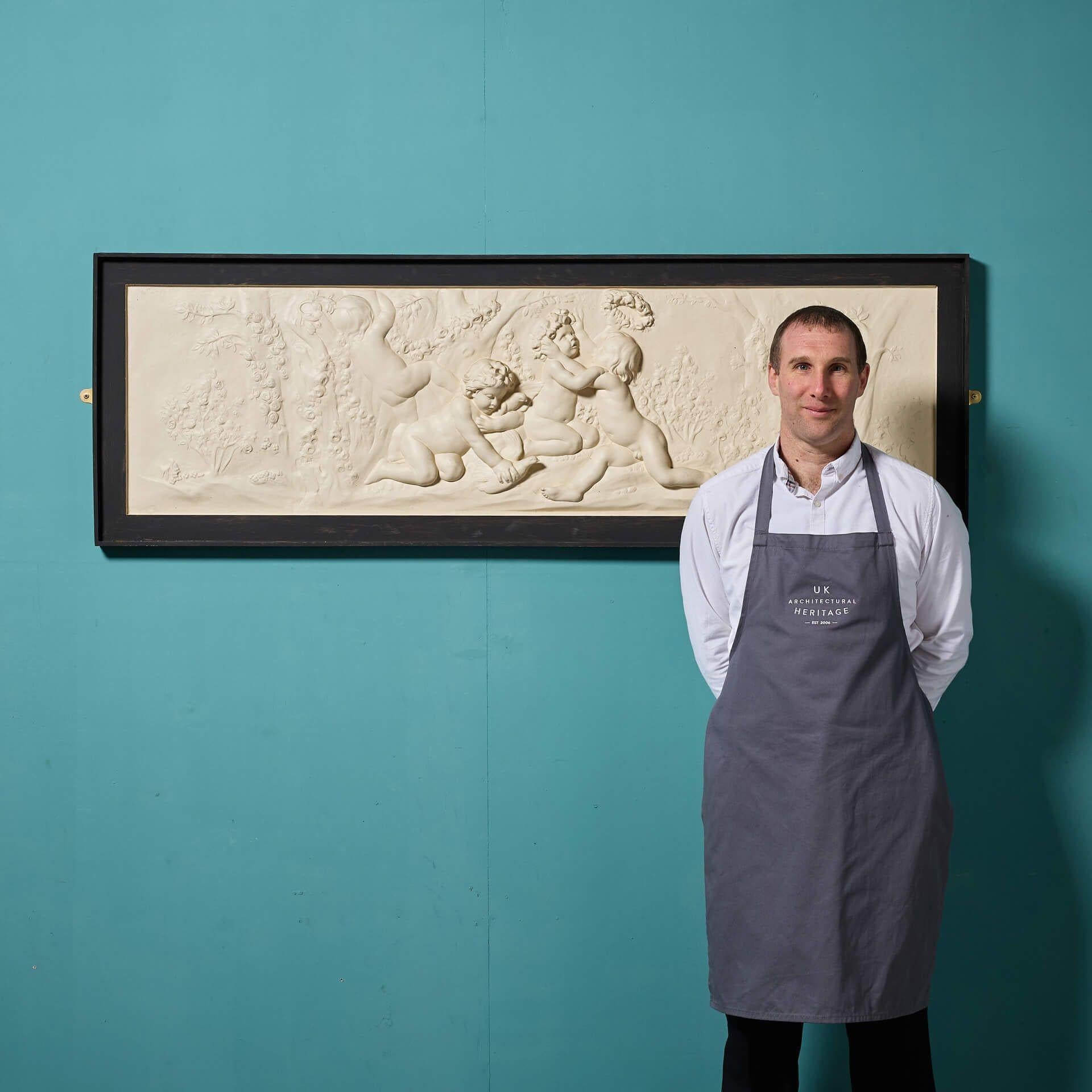 Eine wiederhergestellte Wandtafel aus Gips im neoklassischen Stil mit einem beeindruckenden, mattschwarzen Rahmen.

Diese handgefertigte Plakette in großer Landschaftsform ist mit einem Relief verziert, das eine romantische Szene mit vier