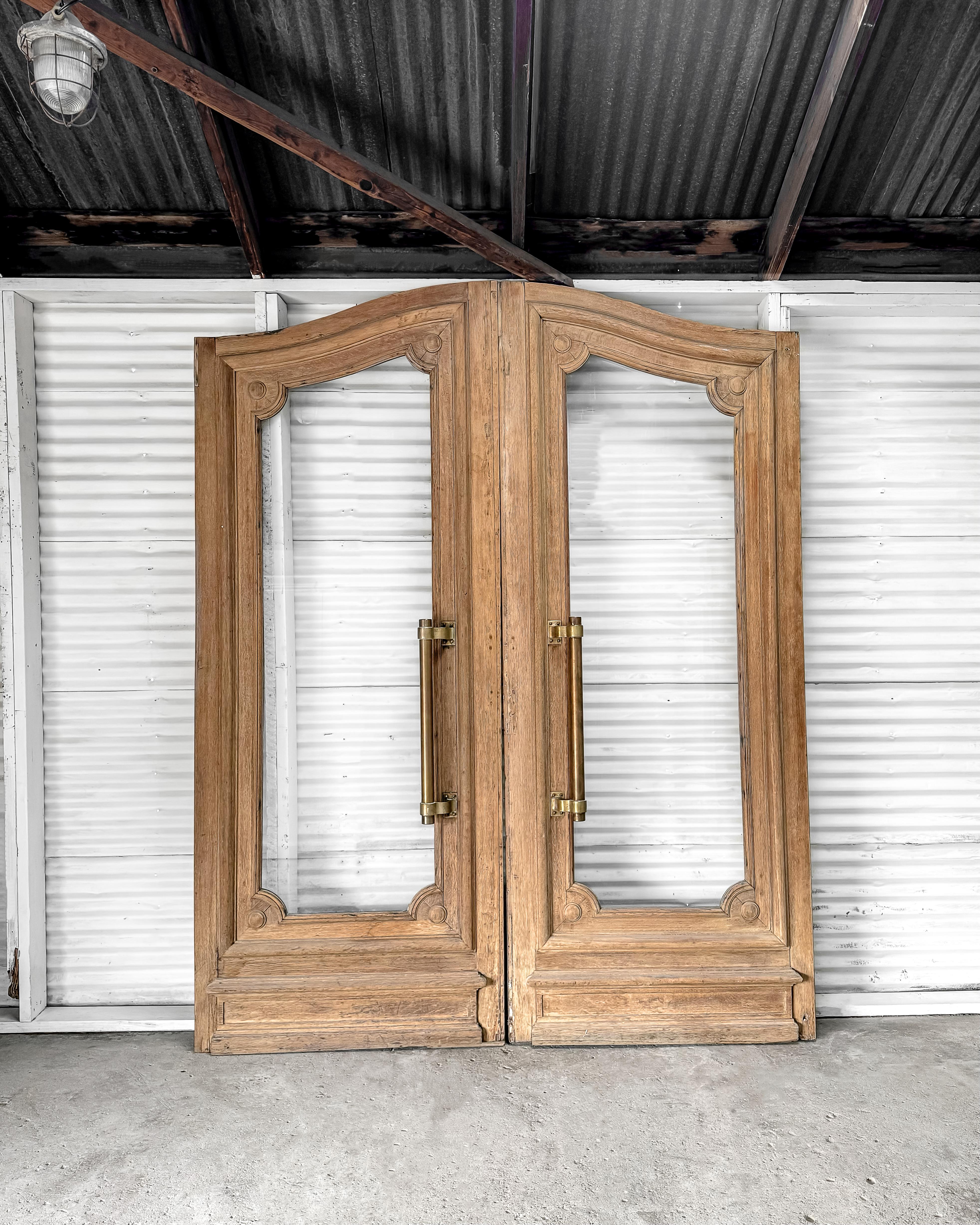 Ein beeindruckendes Paar geborgener Außentüren aus einem Bankgebäude in Frankreich mit einer anmutigen, gewölbten Oberseite und einer natürlichen Holzoberfläche. Jede Tür hat ein einzelnes durchgehendes Fenster, das von einer abgeschrägten Leiste