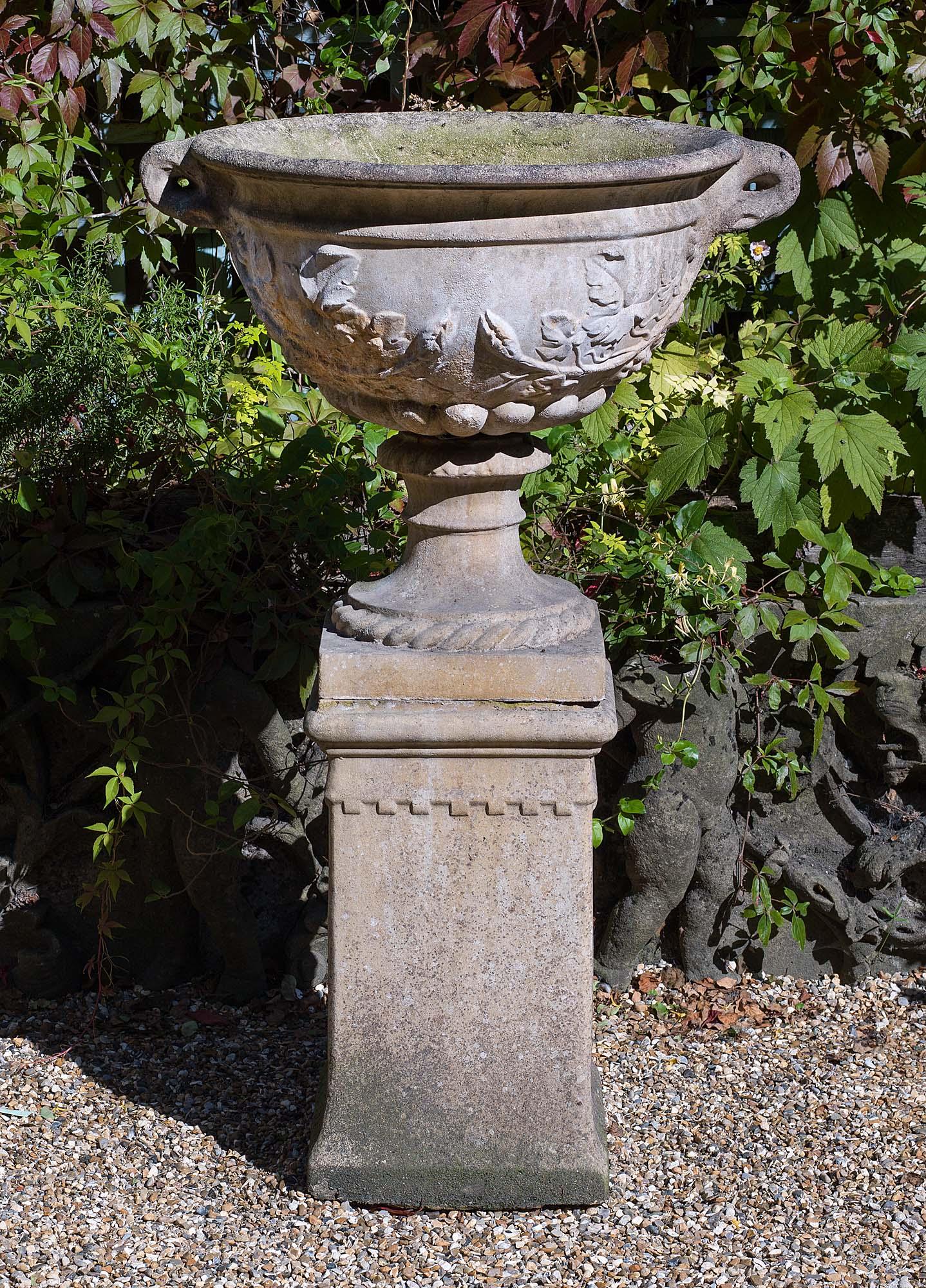 A large reconstituted stone garden urn in the classical tradition. The ovoid body, with carved trailing grape vine decoration, sits on a rope twist socle and is raised on a castellated plinth.
English, mid-20th century.
   