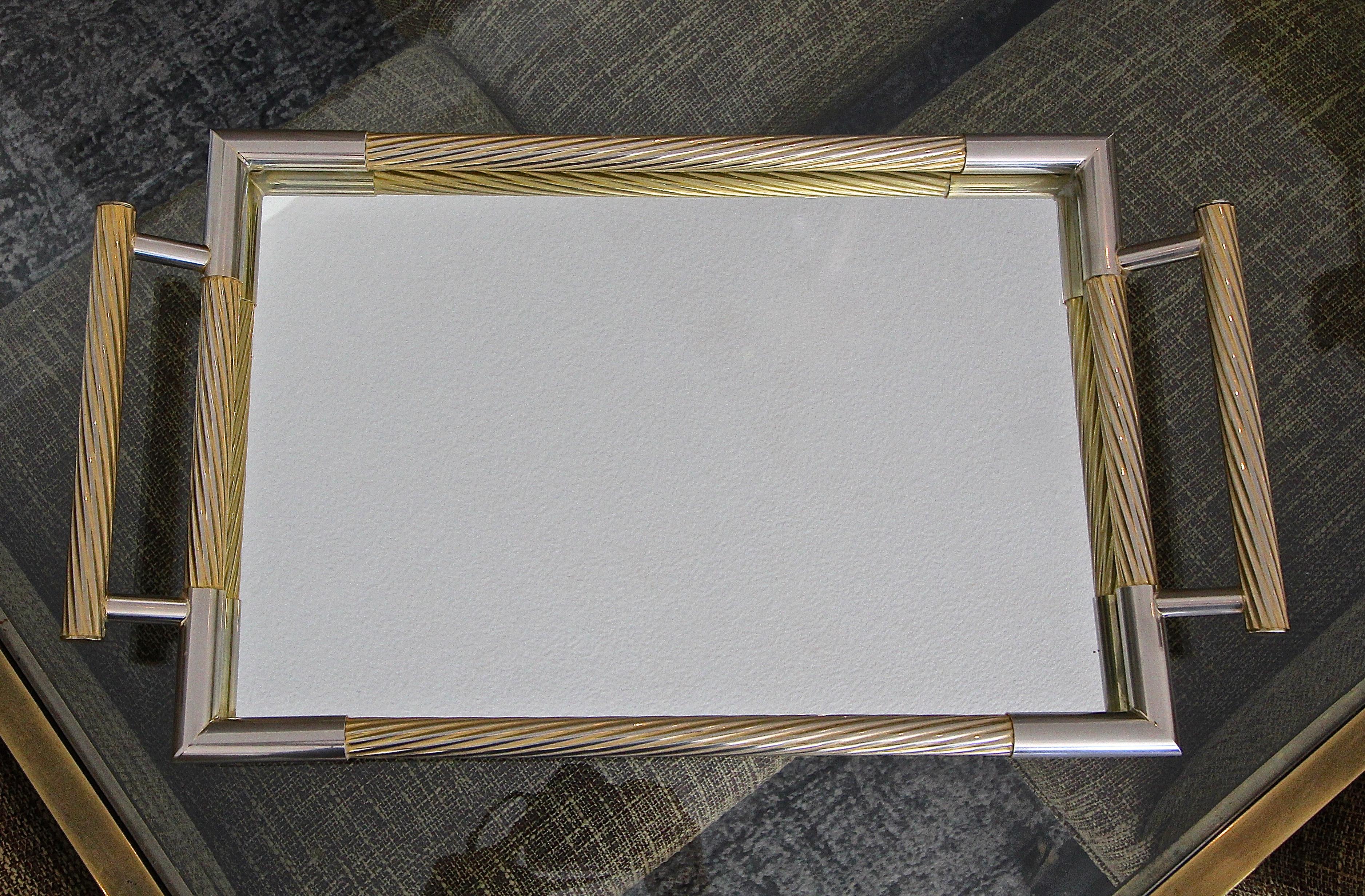 An attractive rectangle shaped serving tray made of non metallic metal with partial gold finish detailing. Inset into the tray is newer mirrored glass. The tray features clean lines with diagonal reeded rails and handles. Length with handles 23