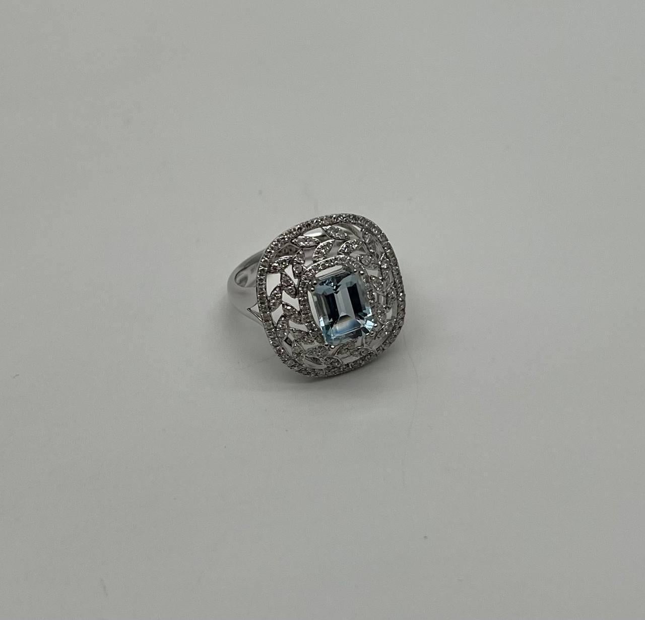This exquisite ring is crafted with intricate detailing. The center features a rectangular blue Aquamarine, surrounded by a halo of accented diamonds in a delicate, ornate pattern. The band is smooth and polished, complementing the intricate design