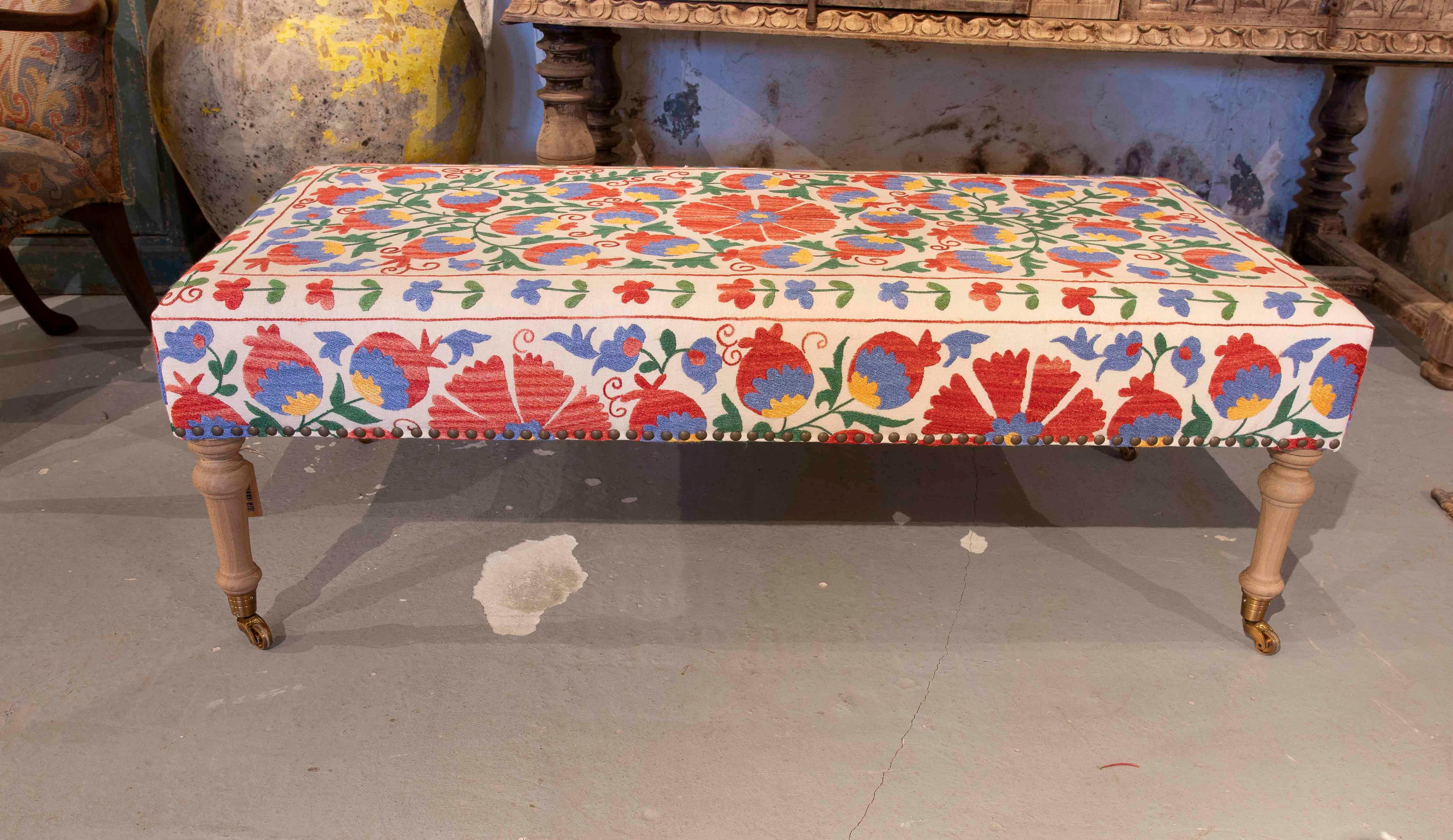 Ce banc rectangulaire est orné d'une tapisserie vibrante, brodée à la main, présentant une explosion de motifs floraux dans des tons de rouge, de bleu, de vert et de jaune. La structure repose sur des pieds tournés en bois clair, complétés par des