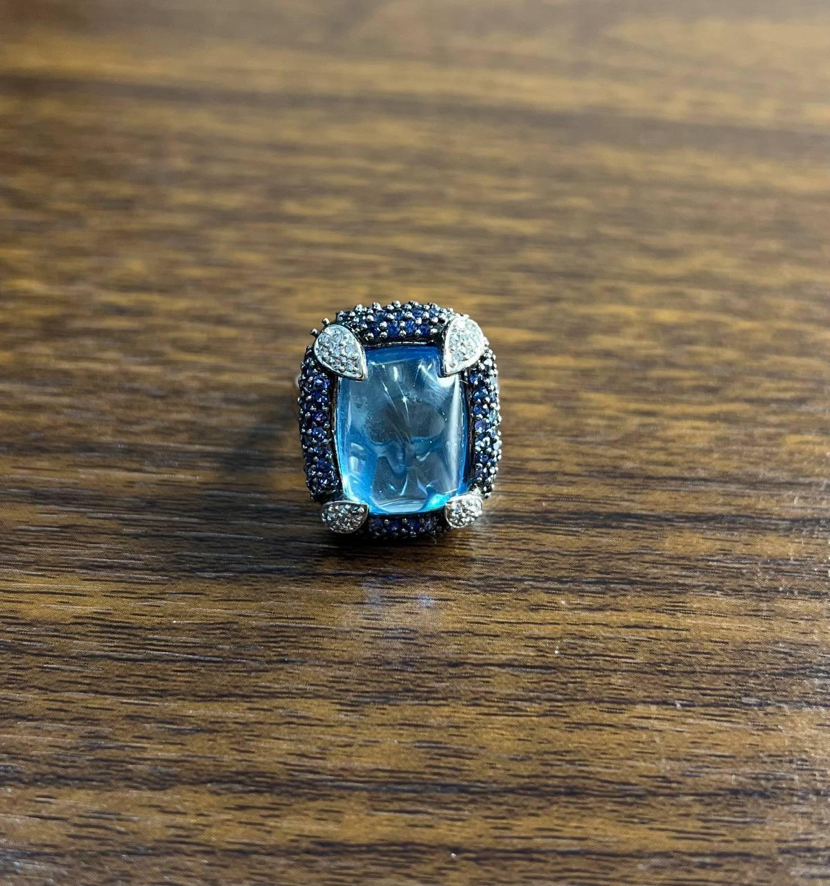 This striking multi-stone ring features a captivating, rectangular blue topaz in sugarloaf cabochon shape at its center. The stone is encased in a halo of deep blue sapphires and white diamonds, adding layers of depth and brilliance. The bold prongs