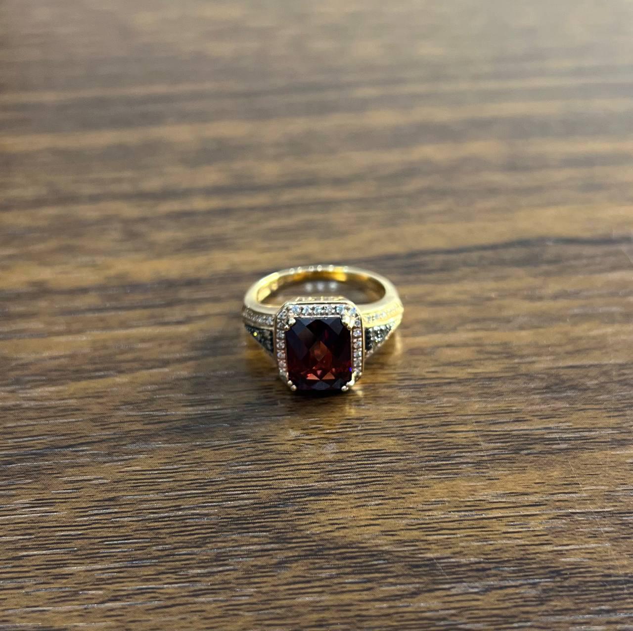 Dieser elegante Ring ist mit einem auffälligen dunkelroten Granat in der Mitte gefertigt. Der Edelstein ist in ein detailliertes 14-karätiges Gold gefasst, das an den Seiten verschlungene Muster aufweist. Der Stein mit quadratischem Schliff ist von