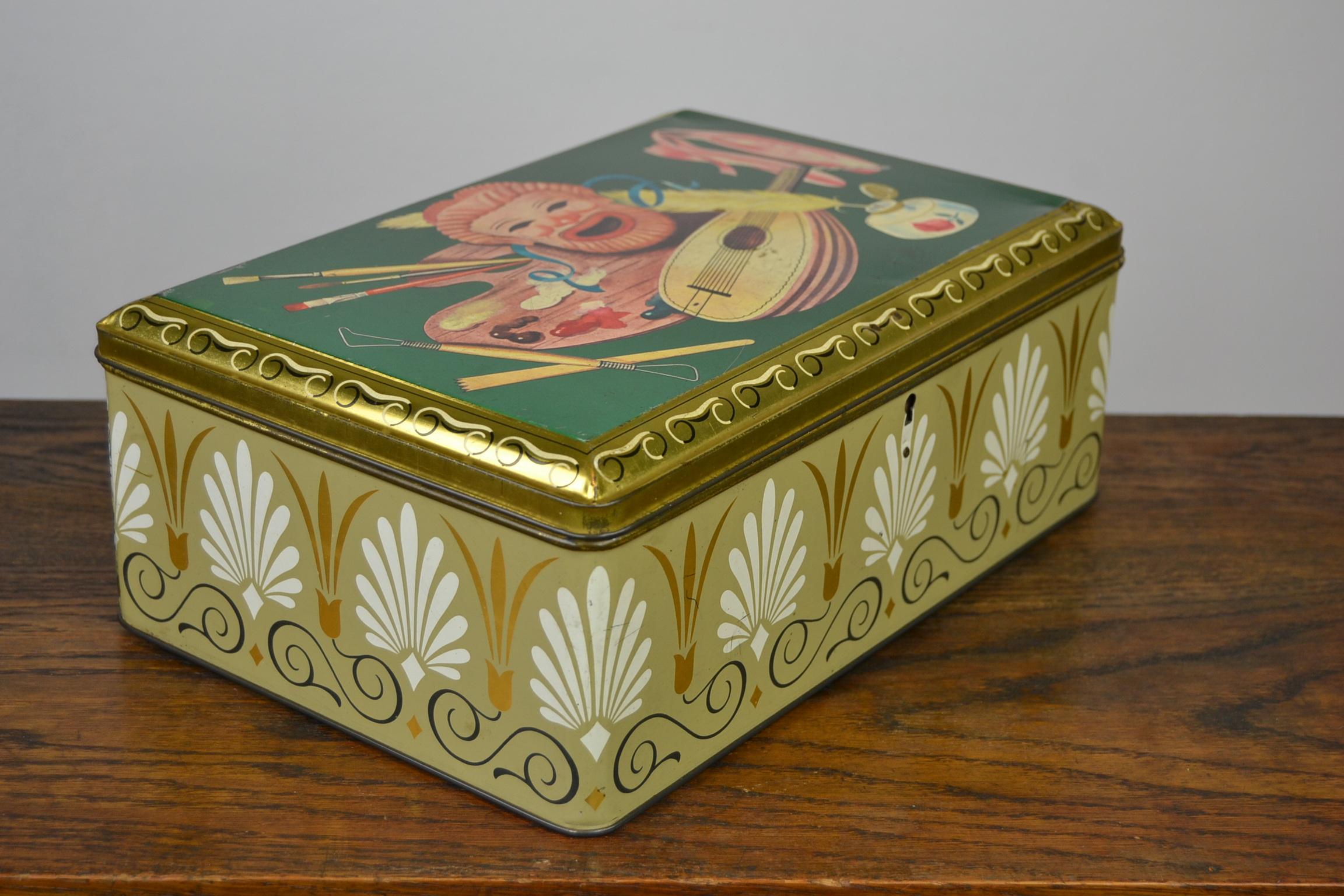 Vintage biscuit tin for Demaret Confectionary, A Belgian Candy Factory.
This vintage tin storage box has a green background with a mask, ballet shoes, inkwell with writing feather, mandoline, painting pallet with brushes and model tools and gold