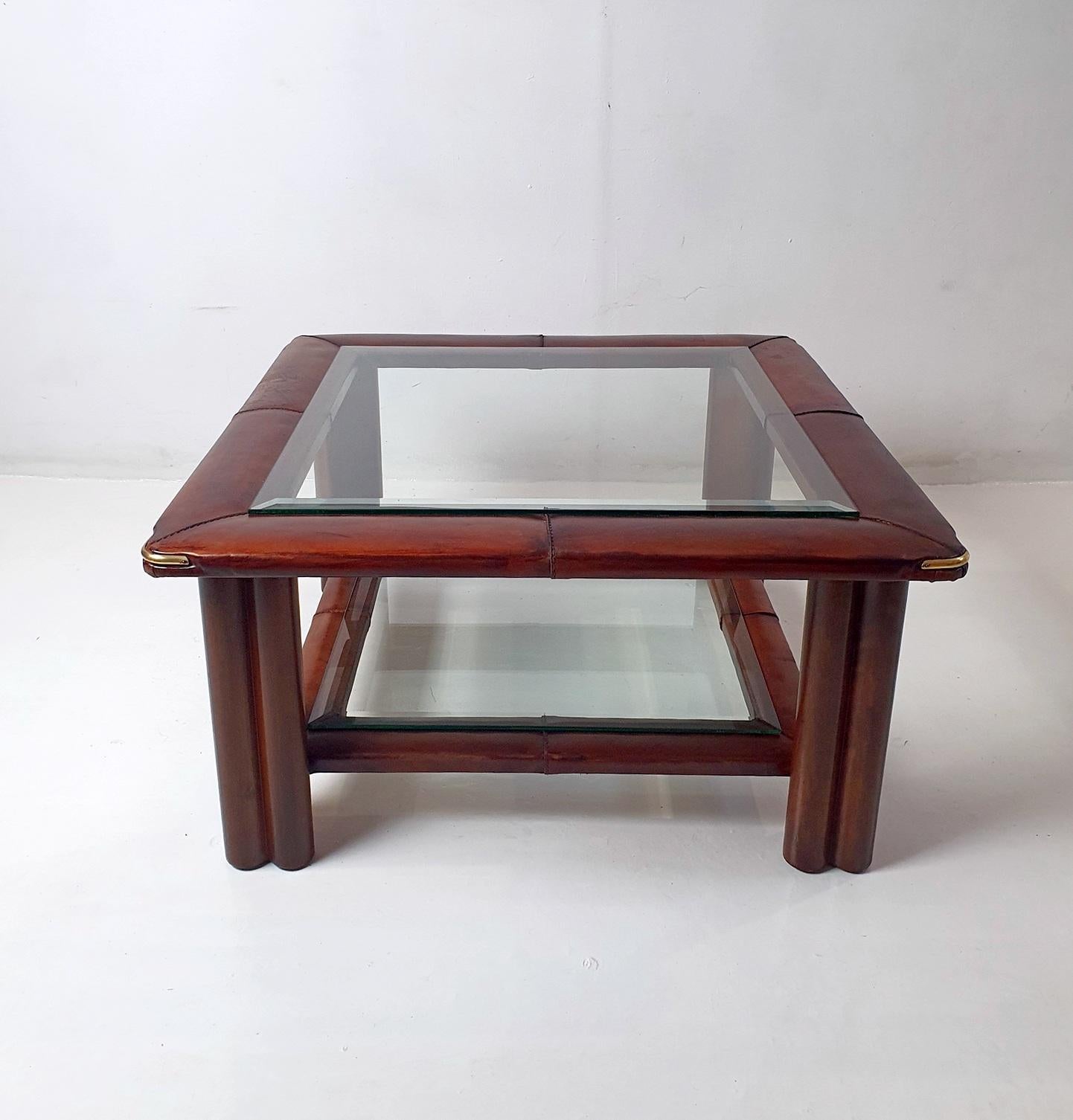 Si tratta di un tavolino da divano prodotto in Italia negli anni '70 in quello che all'epoca era uno stile glam. Realizzato a mano in faggio, pelle, ottone e vetro. Le gambe sono in legno massiccio e le cornici dei piani del tavolo sono circondate