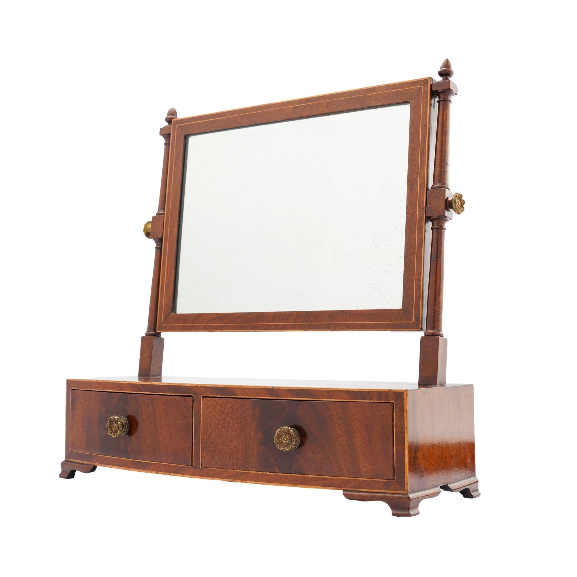 Rectangular mahogany swinger dressing mirror on a bow front stand fitted with a divided drawer. The mirror and stand are inlaid with holly stringing, and the case rests on ogee bracket feet. The drawers are fitted with a single stamped brass rosette