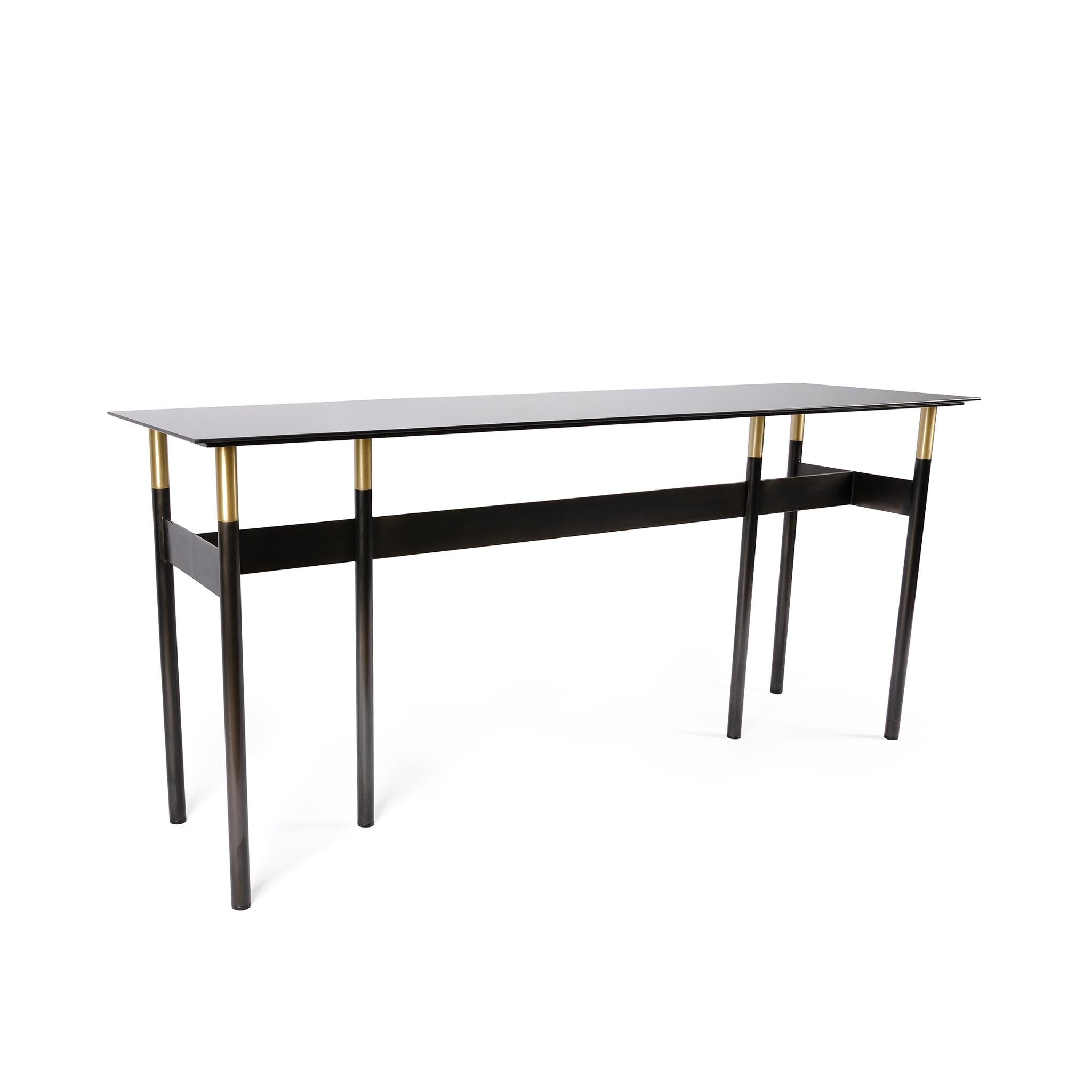 The lank table is rectangular console table with an unassuming elegance. Its dark tones of the linear blackened hand finish steel and tinted mirror glass top contrast against the bright brass details for a unique and sophisticated table. This