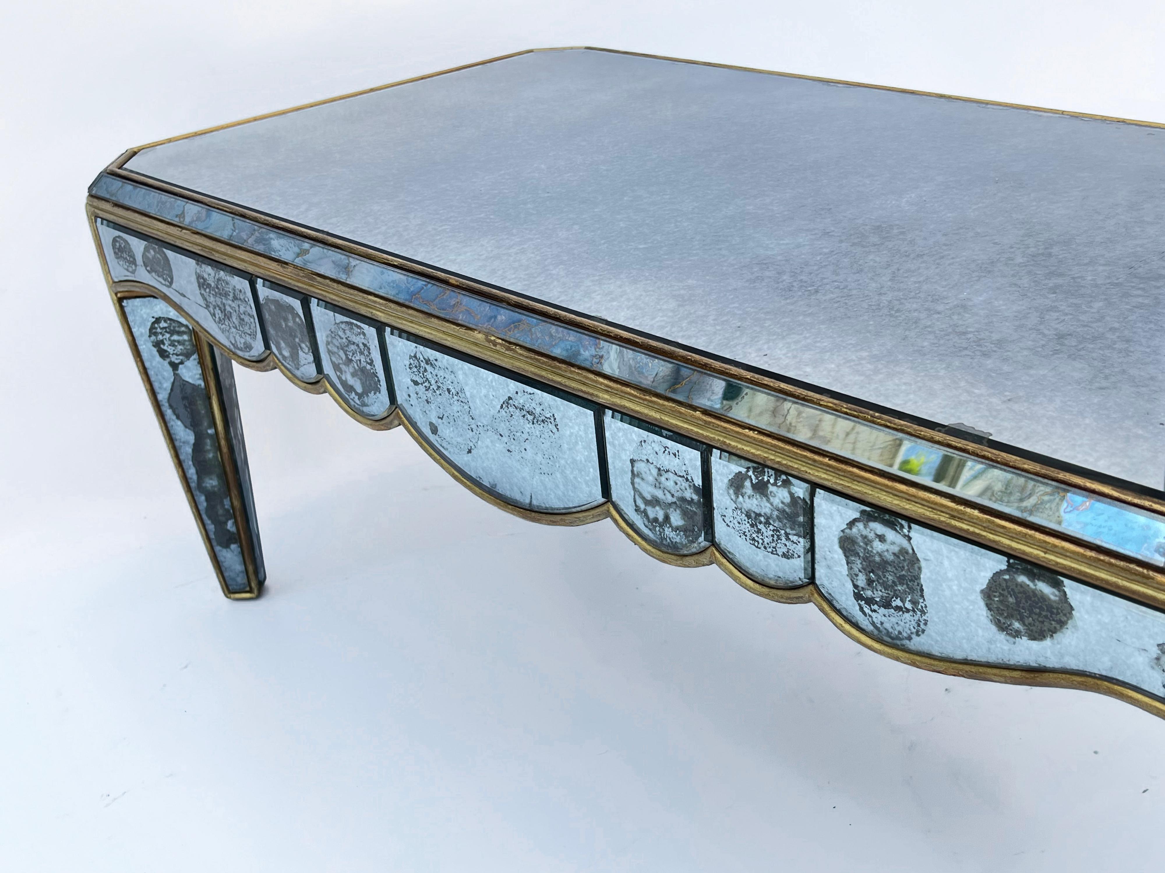 Mid-century coffee table, having a rectangular top with faceted border and canted corners. Its scalloped apron raised on angled, tapering, square legs. All surfaces covered with original, beveled, smoked mirror, now showing desirable loss of
