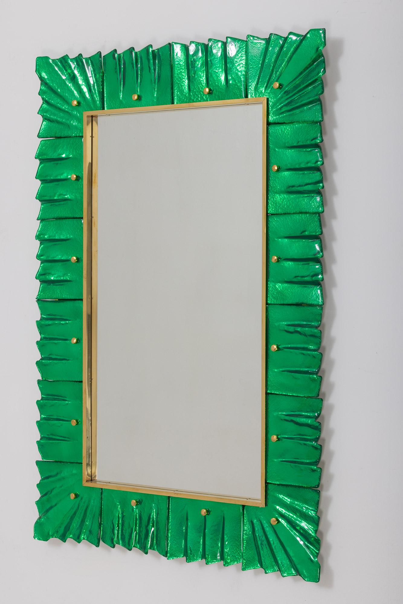 Rectangular contemporary Murano emerald green glass framed mirrors, in stock.
Rectangular mirror plate surrounded with undulating glass tiles in emerald green color held by brass cabochons. 
Handcrafted by a team of artisans in Venice, Italy. 
Can